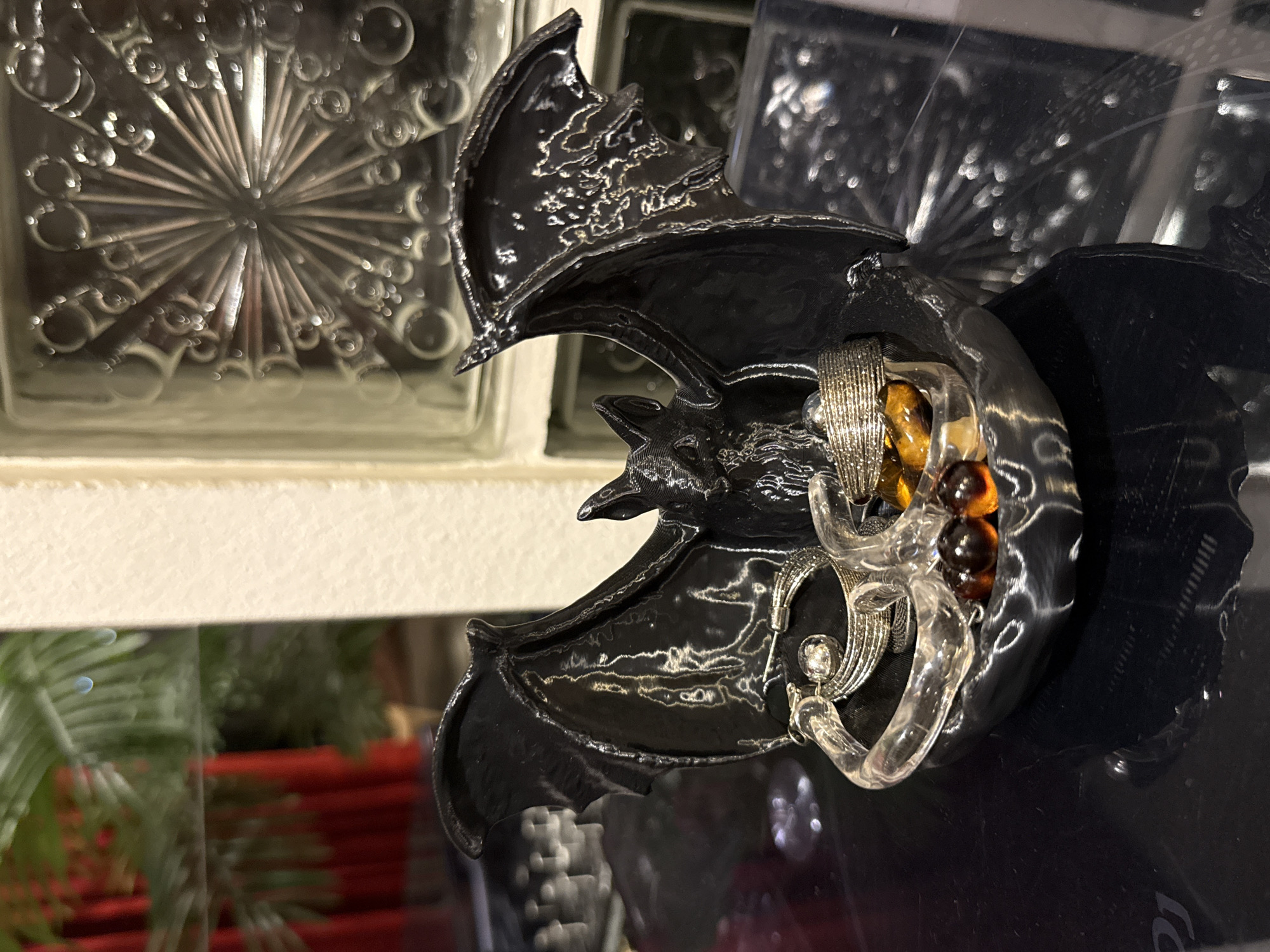 A black, ceramic bat figurine with outstretched wings holds various pieces of jewelry. The bat is positioned on a dark, reflective surface, possibly a table. The jewelry includes several rings, some appearing to be made of silver, and a clear, C-shaped bracelet. There are also a few amber-colored beads. In the background, a frosted glass block wall with a radial pattern is visible. To the left of the bat, through a reflective surface, a hint of green foliage and red drapery can be seen. The overall setting suggests an indoor decorative display. The image was taken at a location in Ko Samui District, Thailand.