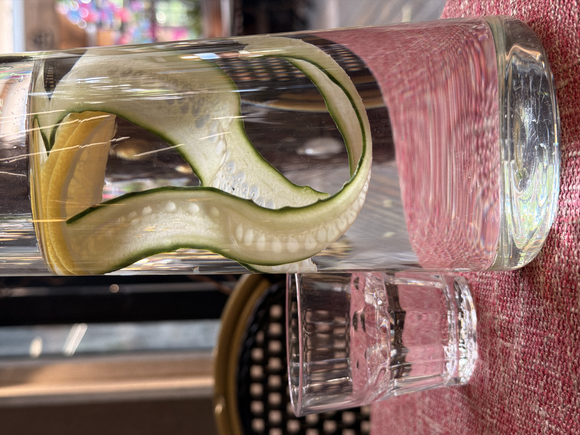 A close-up shot shows a glass pitcher filled with water, sliced lemon, and cucumber ribbons. To the left of the pitcher, a short, clear glass holds water. Both are placed on a textured, reddish-pink tablecloth. In the background, a dark chair with a wicker pattern is visible. The scene appears to be indoors, likely a cafe or restaurant. The image is brightly lit, with a shallow depth of field. The location is not discernible from the image content.