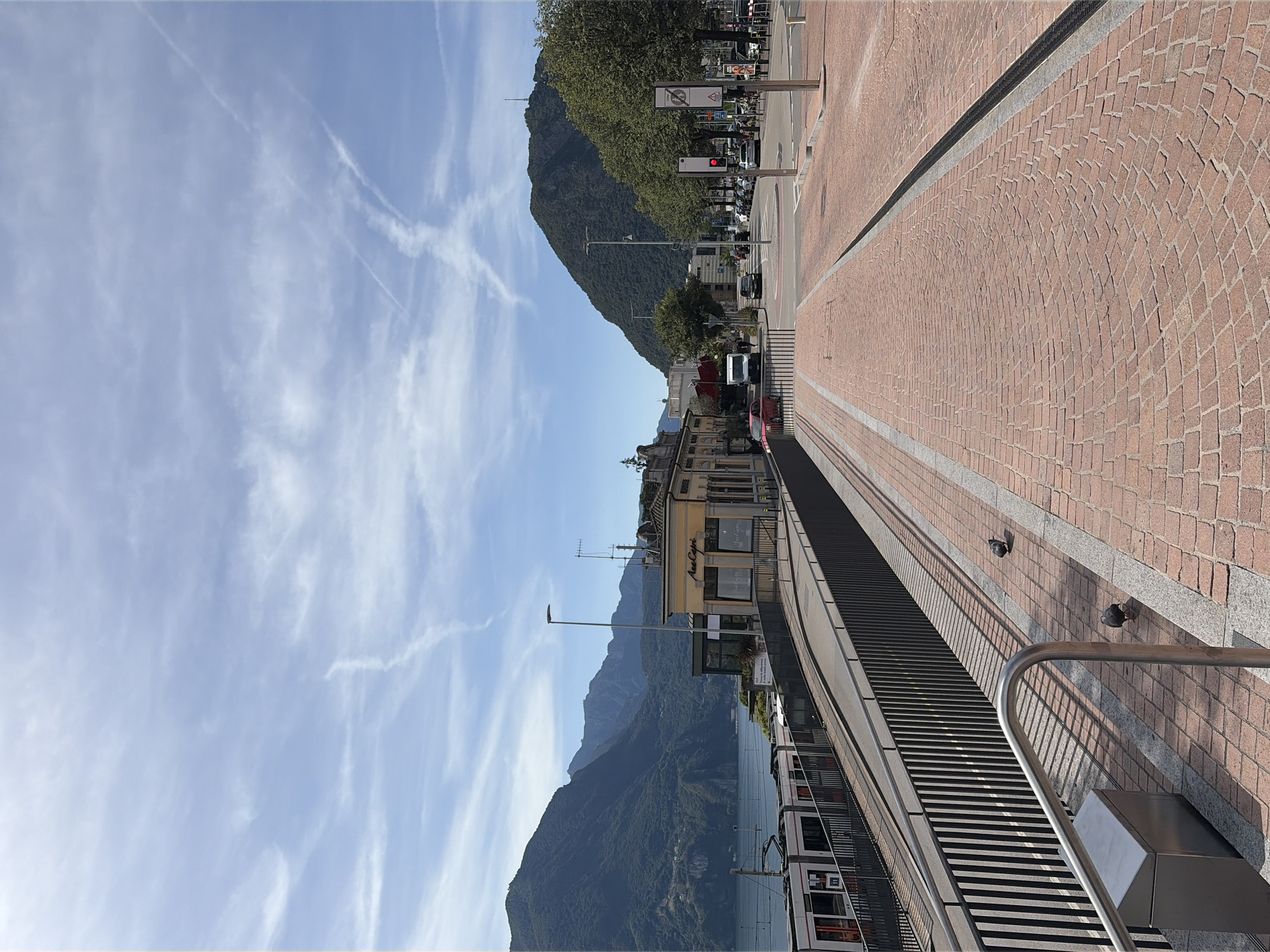 This is a street-level, outdoor, eye-level, medium shot photograph taken in Lugano, Switzerland, on a clear, sunny day. The scene depicts a wide, paved walkway made of red brick, curving towards the right side of the frame. A black metal railing runs along the left edge of the walkway, separating it from a lower level with a tram track and a white tram. The tram is oriented to the left, with its front partially visible. In the background, a body of water, likely Lake Lugano, is visible, flanked by lush, green, tree-covered mountains.
On the right side of the walkway, a tall lamppost stands near a traffic signal that is illuminated red. Further down the street, parked cars line the road, and trees with green foliage create a border. Several buildings are visible, including a yellow building with the text "AmoCapri" on its facade. The sky above is blue with scattered white, wispy clouds, some of which appear to be contrails. Two pigeons are present on the walkway in the foreground. The overall atmosphere is bright and urban, with a prominent natural backdrop.