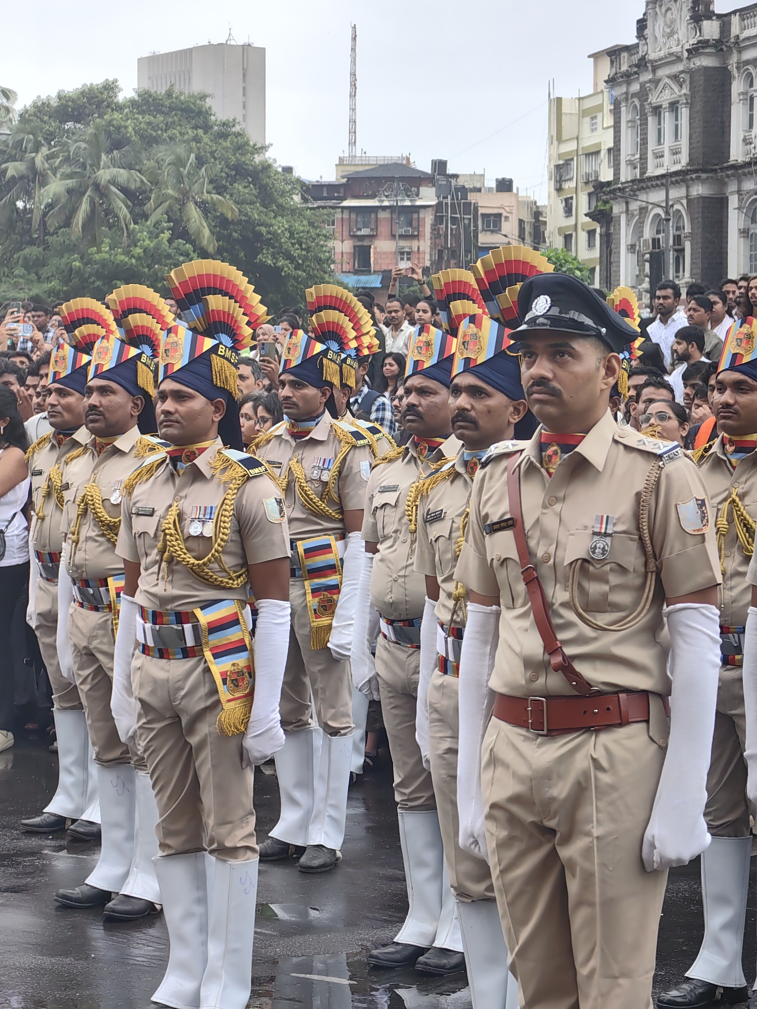 A contingent of police officers stands at attention in the foreground, dressed in tan uniforms with elaborate headdresses featuring red, yellow, and blue plumes, and gold trim. They wear white gloves and tall white boots. The officers are lined up in rows, with some facing directly towards the camera and others at a slight angle. Behind them, a crowd of onlookers is visible, many holding up mobile phones to take pictures. In the background, several buildings are present, including a grand, ornate stone building and a taller, more modern structure. Palm trees are also visible, suggesting a tropical or subtropical climate. The ground appears wet, indicating it may have recently rained or is raining, and there are reflections on the wet surface. The overall scene suggests a public event or parade taking place in Mumbai, India, likely in an area with historical architecture.