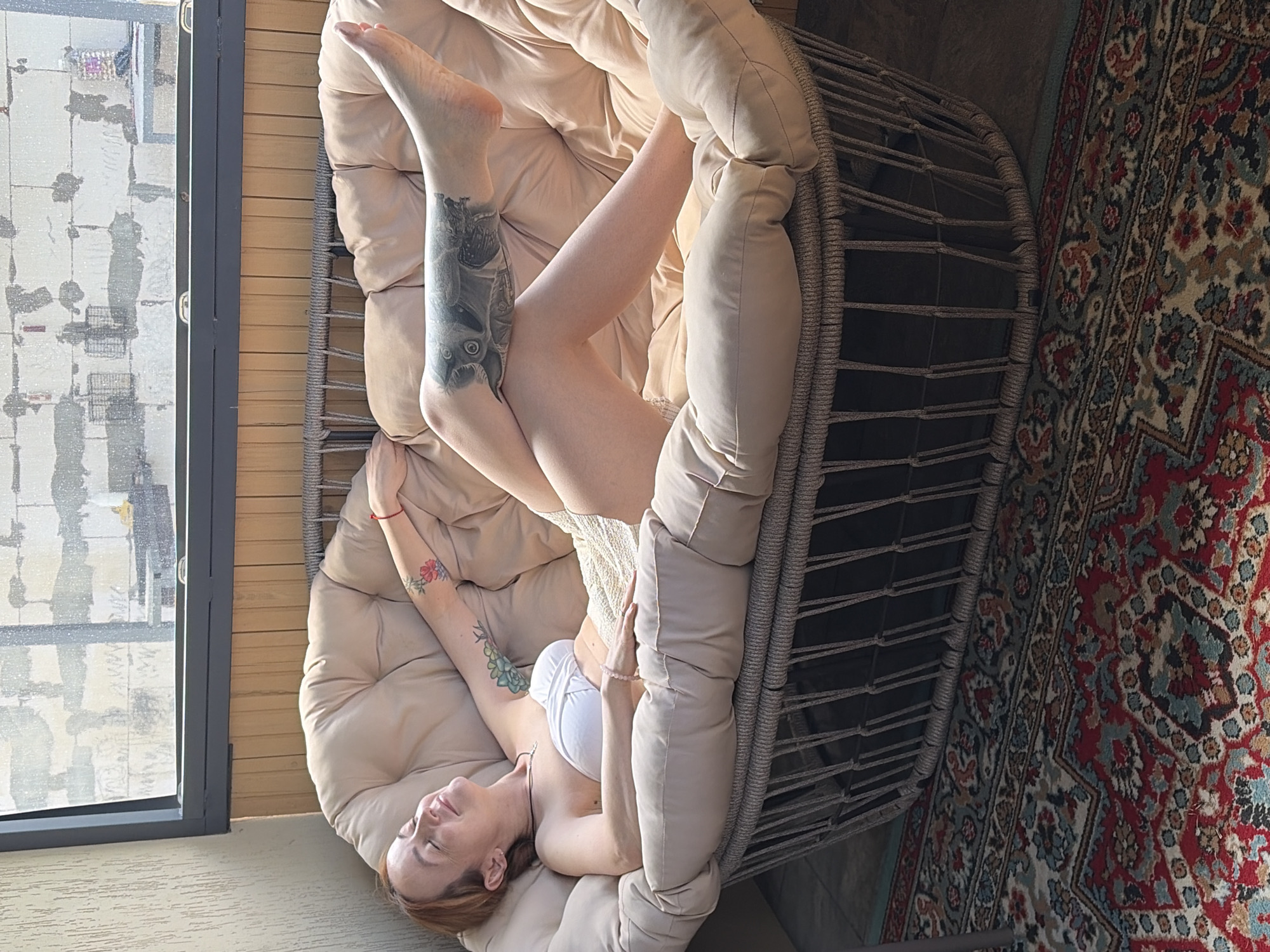 A woman reclines in a papasan chair. She is fair-skinned with red hair. She wears a white bra and a necklace. Her eyes are closed. She has tattoos on both arms and on one leg. The papasan chair is beige with a woven, grey metal frame. The chair sits on a dark floor next to a colorful rug. To the left is a window. The view from the window includes residential buildings. The location is Chaikovsky, Russia.