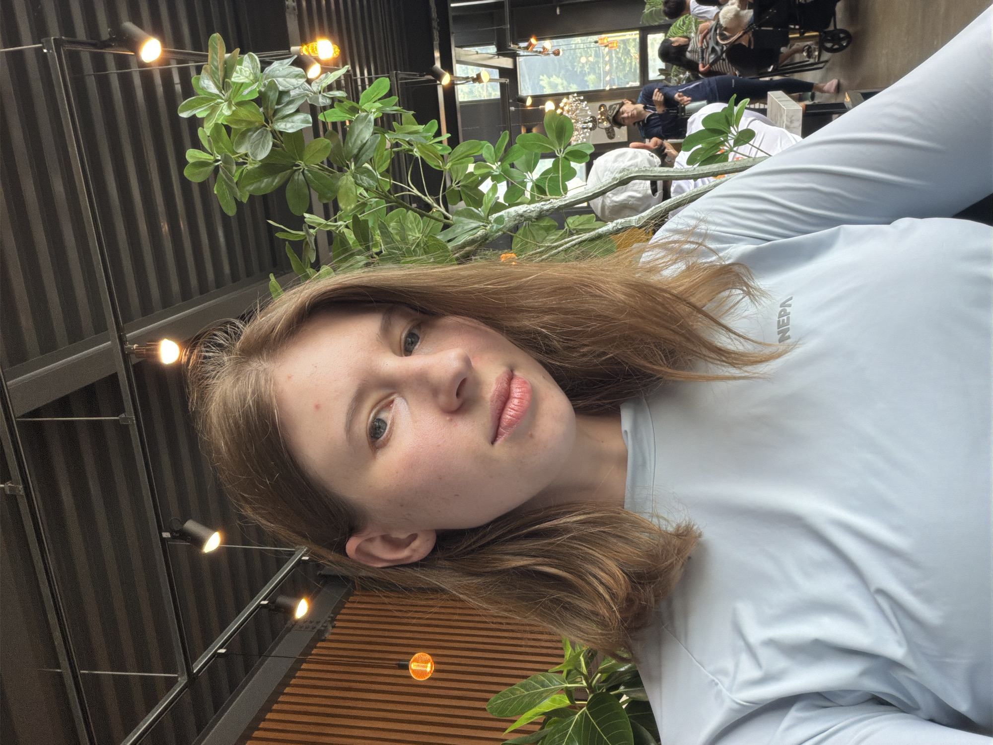 A young Caucasian woman with shoulder-length brown hair and light skin takes a selfie indoors. She is wearing a light blue long-sleeved shirt with the word "NEPA" visible on the chest. The background features an industrial-style ceiling with exposed beams and track lighting, a wooden slat wall, and a large potted plant. In the blurred background, other individuals are seated at tables, suggesting the location is a cafe or restaurant. The scene appears to be in a modern establishment, potentially in Seo-myeon, South Korea.