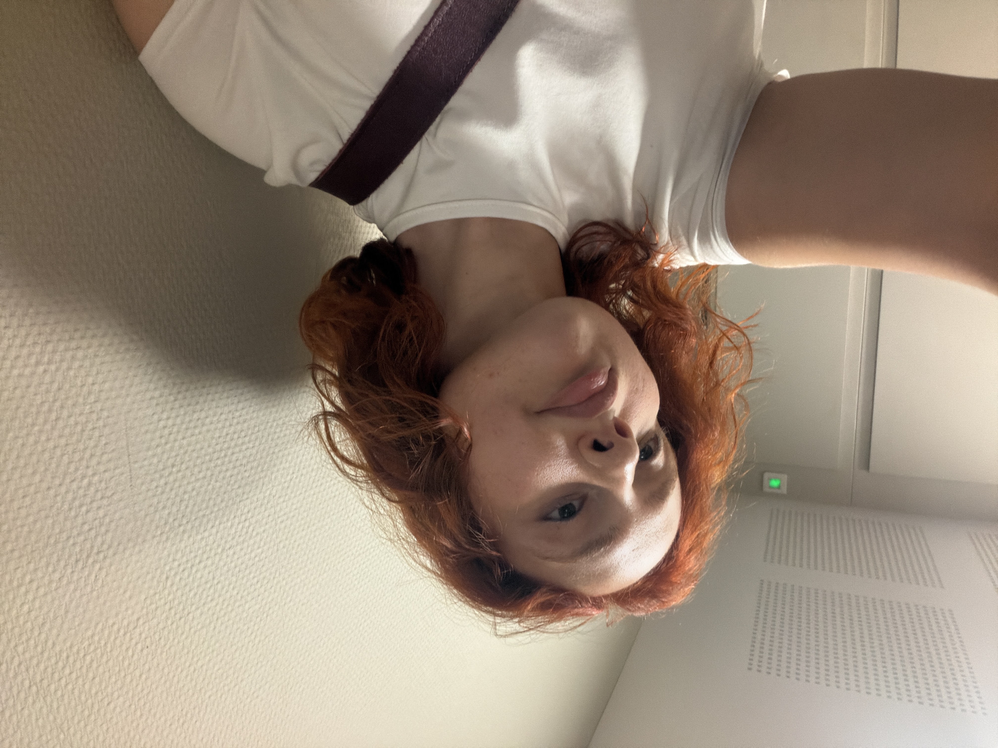 A young woman with vibrant red hair, wearing a white t-shirt and a dark purple strap across her chest, poses for a selfie. She is positioned in what appears to be a hallway or corridor. The background consists of a lightly textured, beige wall and a portion of a white door. Above, a white ceiling with a textured acoustic tile pattern is visible. A small green light is illuminated on the ceiling. The overall lighting suggests an indoor, possibly public or institutional, environment. There is no indication of this location being in Strasbourg, France.