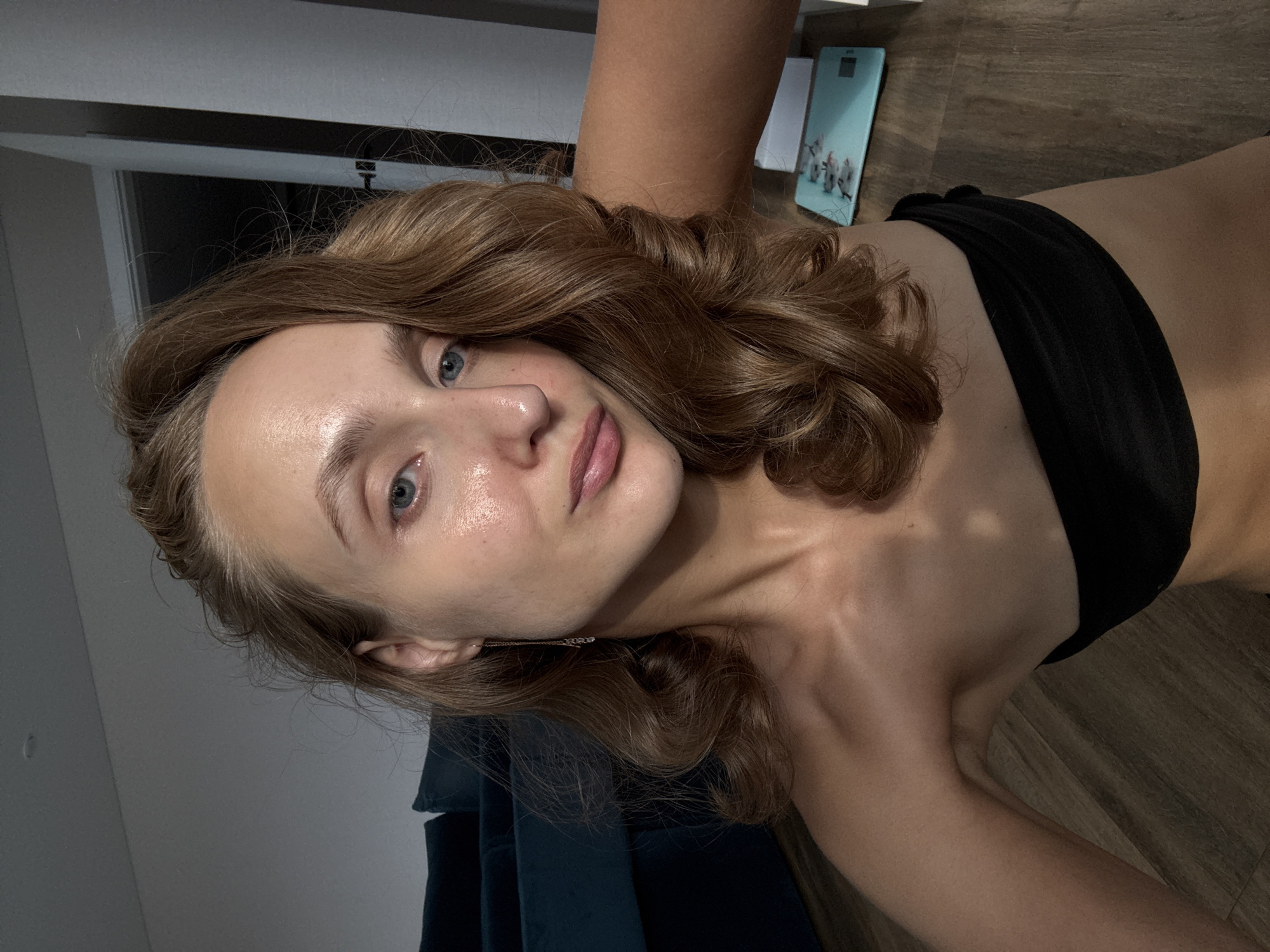 A young woman with light brown wavy hair is photographed from the chest up, looking into the camera. She is wearing a black strapless top. Her skin appears to be glowing, and she has light eye makeup. A delicate gold earring with small charms hangs from her left ear. In the background, a dark teal sofa is partially visible to the left. To the right, a door is ajar, and a light blue scale with a fish pattern is on the floor. The floor is wooden. The scene is indoors, and the lighting suggests it might be evening or a dimly lit room. The location is identified as Podshivalovo, Russia.