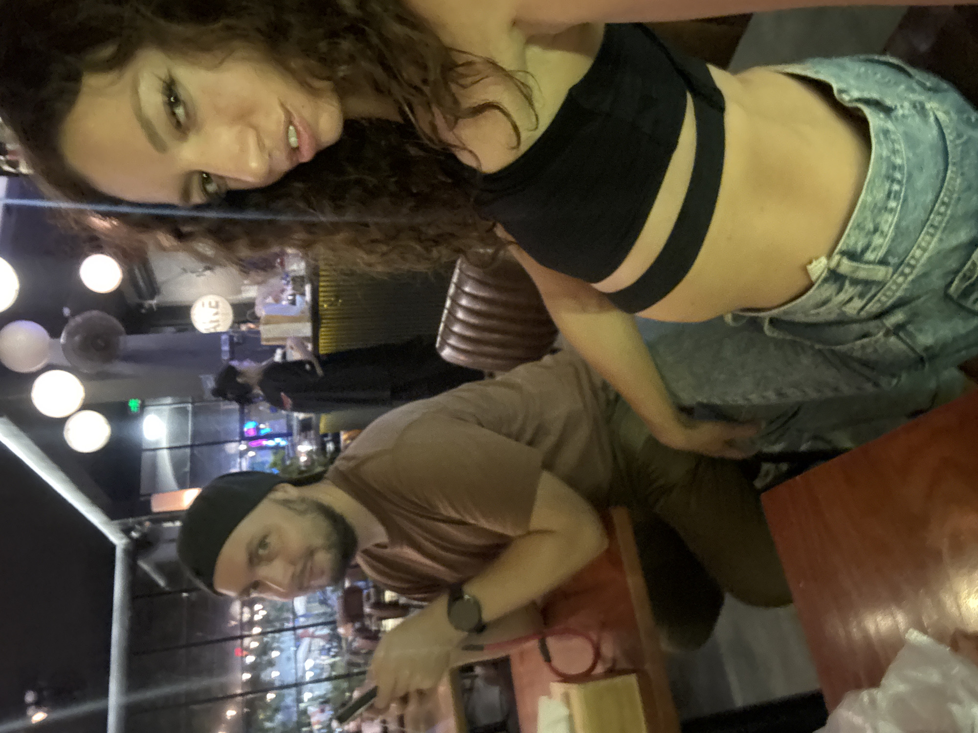 A daytime shot of a man and woman posing for a selfie in Đà Nẵng, Vietnam. The woman is wearing a black bra top and denim shorts, and the man is wearing a brown t-shirt and a black cap. They are seated at an outdoor table with an array of objects including eating utensils. Behind them are lights, people, and a cityscape.