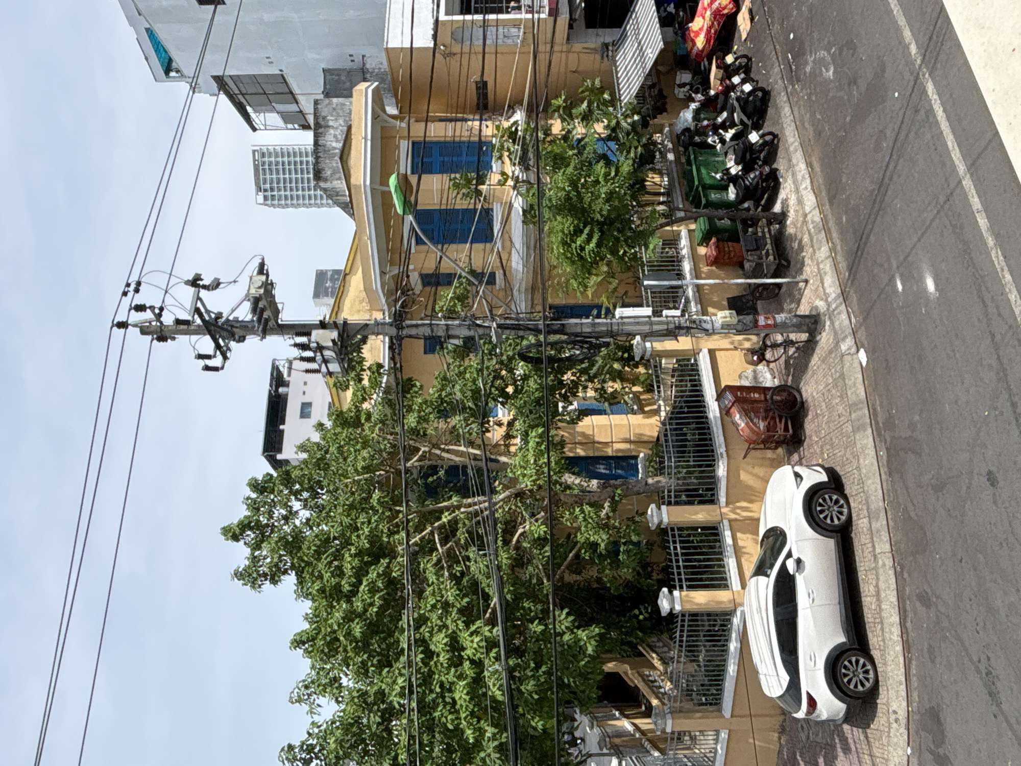 This image captures a street scene in Nha Trang Ward, Vietnam. A white sedan is parked on the right side of a paved road, its wheels turned slightly. To the left of the road, a yellow building with blue-shuttered windows stands behind a metal fence. A large green tree with dense foliage is prominently featured in the foreground, its branches intertwining with a complex network of overhead electrical wires. In the background, several other buildings of varying heights and architectural styles are visible, including a modern high-rise structure in the distance. Several black motorcycles are parked near the yellow building, alongside green trash bins and a red cart. The scene appears to be set during the daytime, with shadows indicating the sun's position. The sky is overcast, suggesting cloudy weather. The presence of numerous electrical wires and the density of the urban environment are characteristic of many streets in Vietnam. There is no visible text in the image.