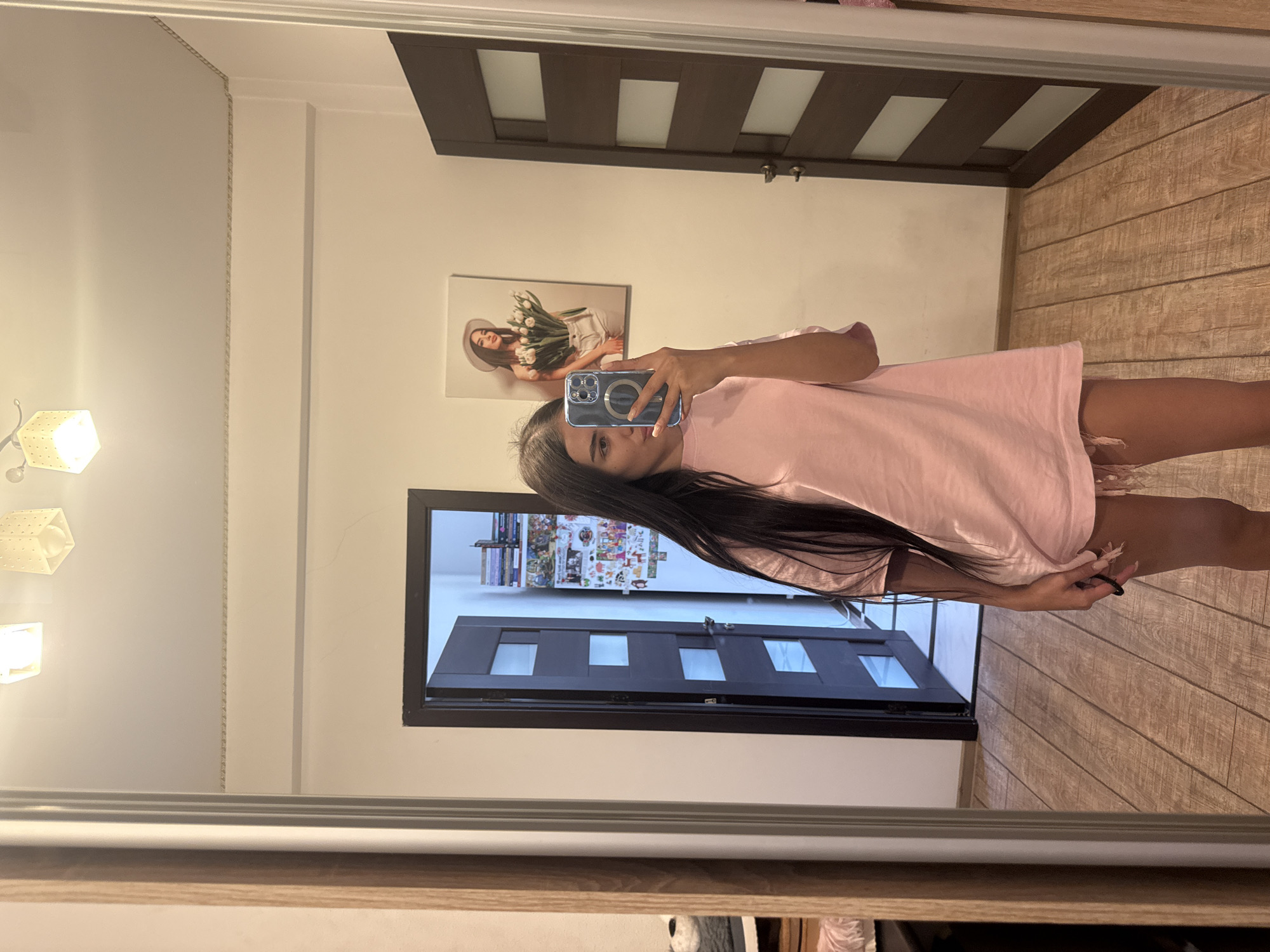A young woman with long, dark hair is standing in a room, taking a selfie in a large mirror. She is wearing a pink t-shirt that extends to her mid-thighs. The room has wooden floors and white walls. There is a closed dark wooden door with frosted glass panels to her right. To her left, a closet with a wooden frame is partially visible, with some items on shelves inside. On the wall to her left, a framed picture shows a woman holding a bouquet of tulips. Another framed picture is partially visible above it, showing a woman's face. A ceiling light fixture with multiple square lamps is visible at the top of the frame. The room appears to be a bedroom or dressing area.