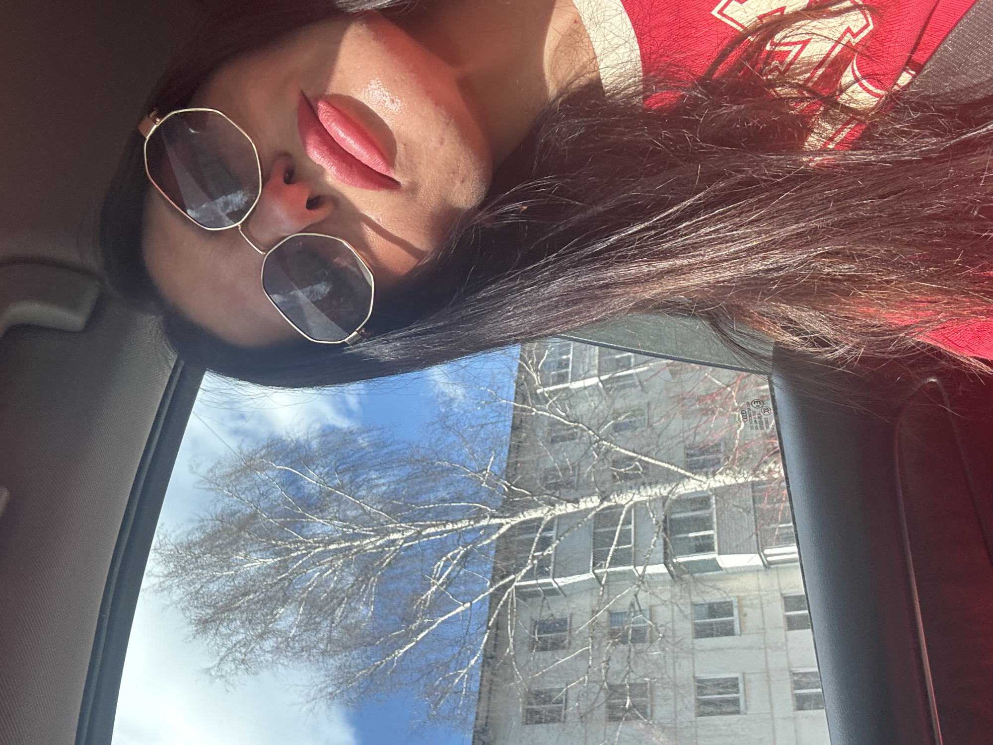 A selfie photo taken from inside a car shows a woman wearing sunglasses and a red shirt with what appears to be a sports team logo. She is positioned in the upper part of the frame. The view outside the car window reveals a tall, leafless tree against a blue sky, with a multi-story building partially visible behind the tree. Based on the architecture and context clues, the building is consistent with residential architecture from Chaikovsky, Russia. The time of year depicted seems to be late winter or early spring, as indicated by the bare tree.