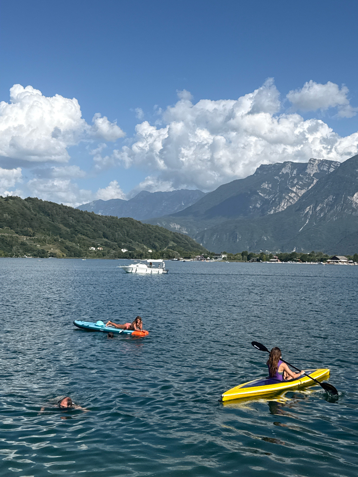In the city of Calceranica al Lago, Italy, on a clear day with scattered clouds, a vast, calm body of water reflects the sky. In the foreground, a person wearing a life vest is swimming. Further out, a woman lies on a blue and orange paddleboard, while another woman in a purple swimsuit rows a yellow kayak with a black paddle. A white motorboat travels in the middle ground, and in the background, lush green hills rise to meet majestic, rocky mountains under a blue sky.