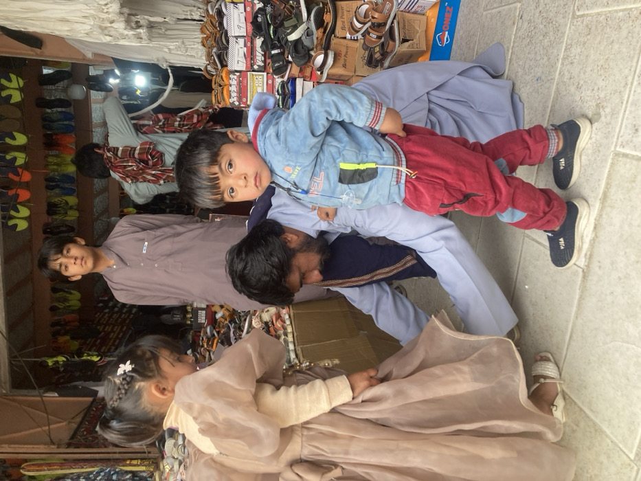 This image captures a scene within a market stall in Taftan, Pakistan. In the immediate foreground, a toddler boy stands facing the viewer, dressed in a light blue hooded jacket with yellow and black details, red pants, and dark blue sneakers. A white pacifier is clipped to his jacket. To his left, an adult male with a dark beard and light blue shalwar kameez is crouched, seemingly attending to the footwear or lower attire of a young girl. The girl, positioned to the left of the crouching man, wears a light brown dress with voluminous sheer sleeves and white sandals. Her dark hair is styled in pigtails, with a white floral clip visible on the right pigtail. She is looking downwards. In the background, a teenage boy stands upright, wearing a light brown shalwar kameez, observing the scene. Further back and to the right, another adult male, partially obscured, is visible from the back, wearing light clothing and a reddish-brown patterned scarf draped over his shoulder, appearing to interact with merchandise.
The stall itself is a footwear vendor, evidenced by numerous pairs of shoes, sandals, and slippers displayed on various surfaces. Brightly colored slippers hang from a wall in the background, while stacks of shoe boxes, some labeled "Best Power," are visible on the right, topped with assorted sandals. Piles of footwear are also present on a low display surface to the left of the crouching man and girl. A fluorescent light tube is mounted in the background. The floor is covered with light-colored square tiles.