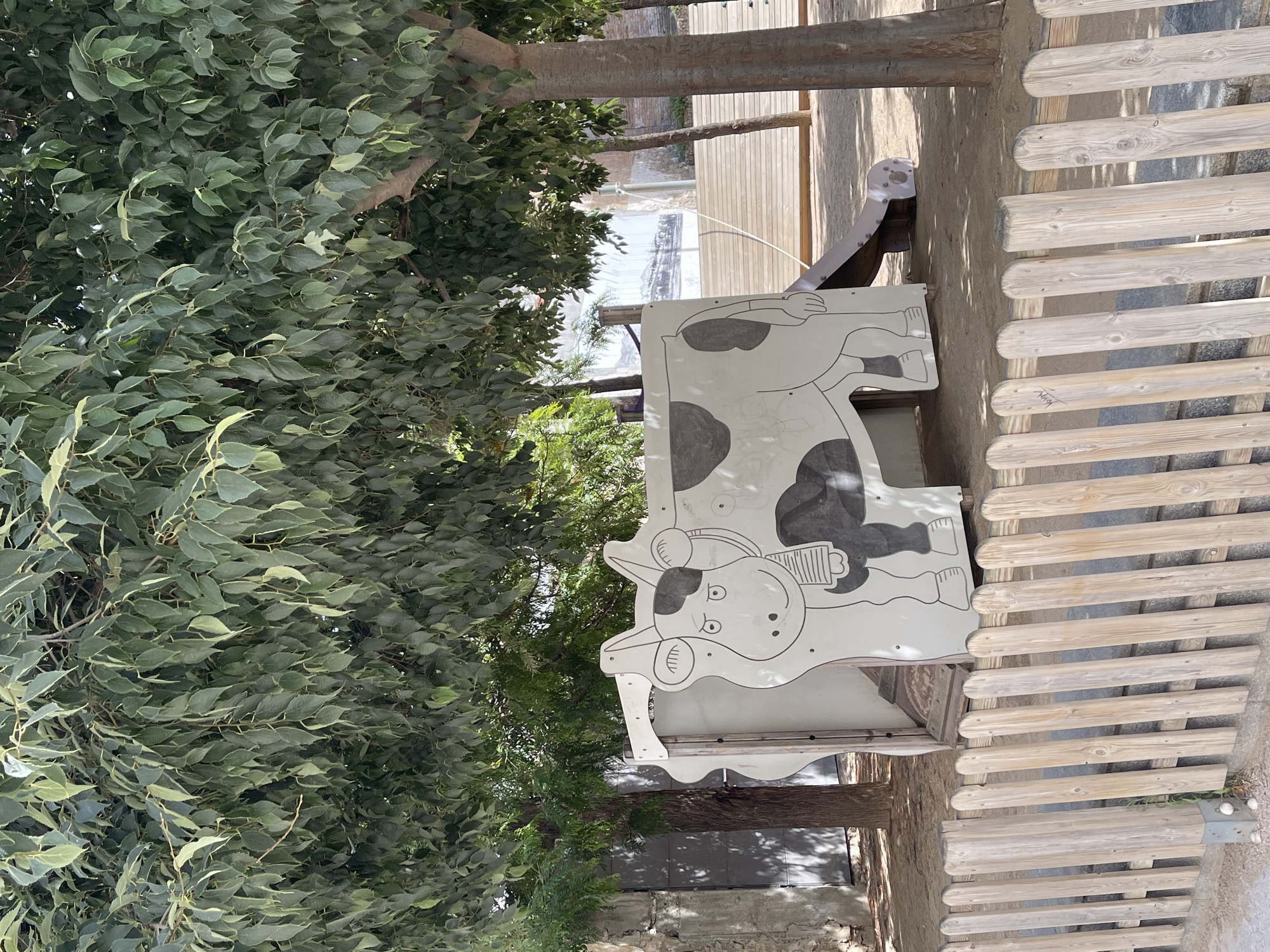 The image captures a playful outdoor scene in Santa Coloma de Gramenet, Spain, featuring a whimsical playground structure. In the foreground, a rustic wooden fence made of vertical planks with rounded tops encloses a sandy area. Beyond the fence, a large, stylized cow-shaped playhouse dominates the scene. The cow is depicted in black and white with simple cartoon-like features, including spots, a bell around its neck, and a friendly expression. A small slide emerges from the side of the playhouse, curving downwards towards the ground.
The environment is shaded by the lush green foliage of mature trees, their branches and leaves creating a dappled sunlight effect on the ground. The ground itself appears to be a mixture of sand and dirt, characteristic of a park or playground. In the background, hints of urban infrastructure can be seen, including a concrete wall and a large, gray metal door on the left, and a wooden elevated platform with stairs on the right. The overall atmosphere is one of a serene, sun-dappled playground, likely a place for children to enjoy outdoor activities.