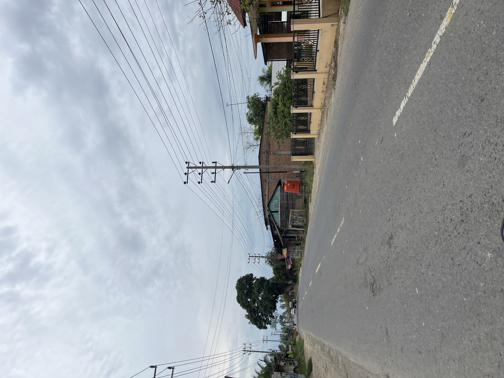 The image depicts a street scene in Simpangempat, Indonesia, under an overcast sky. The paved road, marked with faded yellow lines, stretches into the distance, with a slight curve to the left. Several utility poles with numerous power lines are visible on both sides of the road, some carrying substantial bundles of wires.
On the right side of the road, there is a beige wall topped with a black metal fence, enclosing properties. Behind the fence, a multi-story building with an orange-tiled roof and light-colored walls is partially visible. Further down the road on this side, a single-story building with a dark blue-green roof and wooden siding is situated, with an orange water tank next to it.
On the left side of the road, a row of low-rise buildings with various roofing materials and awnings lines the street. A large, mature tree with a dense canopy stands prominently in the middle ground on the left, its branches extending over the road. Several vehicles, including at least two cars and what appears to be a three-wheeled vehicle, are traveling on the road in the distance. Palm trees and other vegetation are scattered throughout the scene. The overall impression is of a rural or semi-urban environment with active infrastructure and residential or commercial structures.