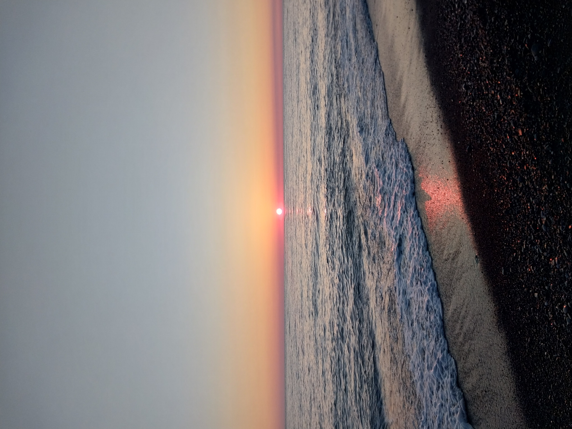 This vertical photograph captures a serene sunrise or sunset over the sea, likely the Black Sea in Sochi, Russia. The image is divided by a distinct horizon line running horizontally across its middle, with the sky on the left and the ocean and shoreline on the right.
The sky, occupying the left half of the frame, transitions gradually from a soft, pale blue-grey at the top left to warm hues of orange and pink closer to the horizon. A clear, bright red-orange sun, visible as a distinct disc, is positioned very low on the horizon, slightly left of the image's vertical center.
The right side of the image features the ocean, with gentle waves reflecting the warm light of the sun. The water exhibits a range of cool tones, from grey to muted blue, interspersed with orange and pink reflections. Towards the far right, the dark, wet beach, possibly composed of sand and pebbles, comes into view. The retreating waves leave a foamy white edge where they meet the shore, and patches of the wet sand also reflect the sun's vibrant light, creating a glimmering effect.
There are no people, man-made objects, or visible text in the photograph. The scene evokes a sense of tranquility and natural beauty, characteristic of a peaceful morning or evening by the sea. The weather appears clear and calm.