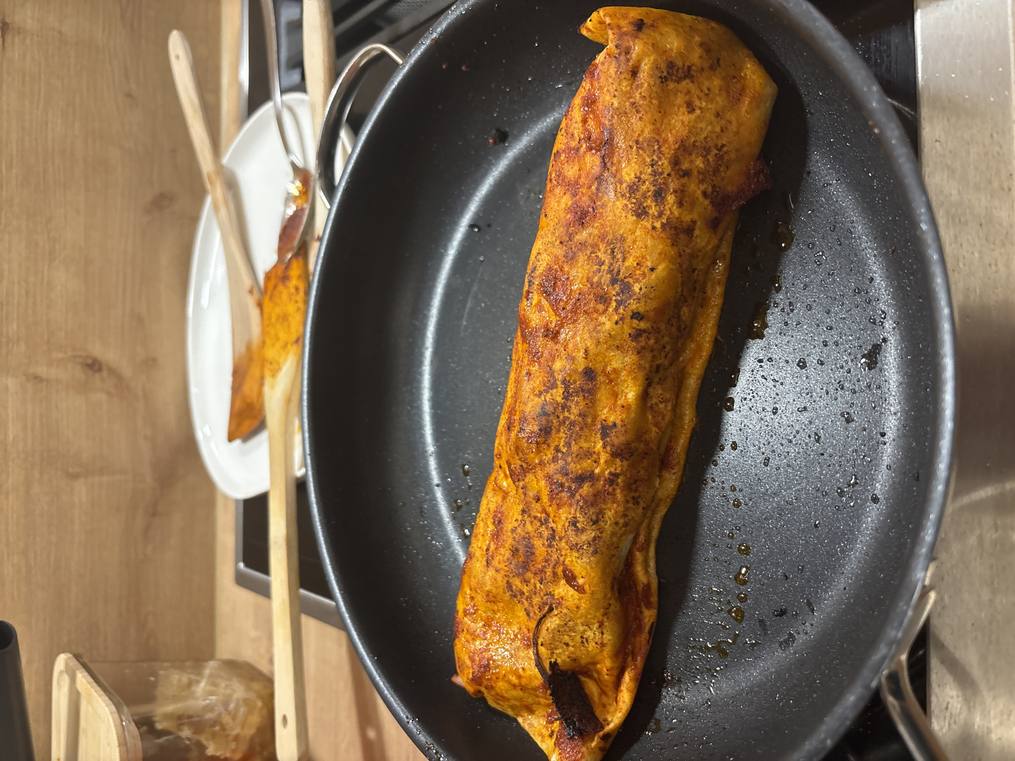 An uncooked enchilada is shown in a frying pan on a stovetop in a kitchen in Viernheim, Germany. The enchilada is golden brown and has red sauce on it. There is some oil in the pan. In the background, there is a wooden counter and a wooden backsplash. There is also a white plate with some red sauce on it and two wooden spoons. A glass jar with a wooden lid is also visible.