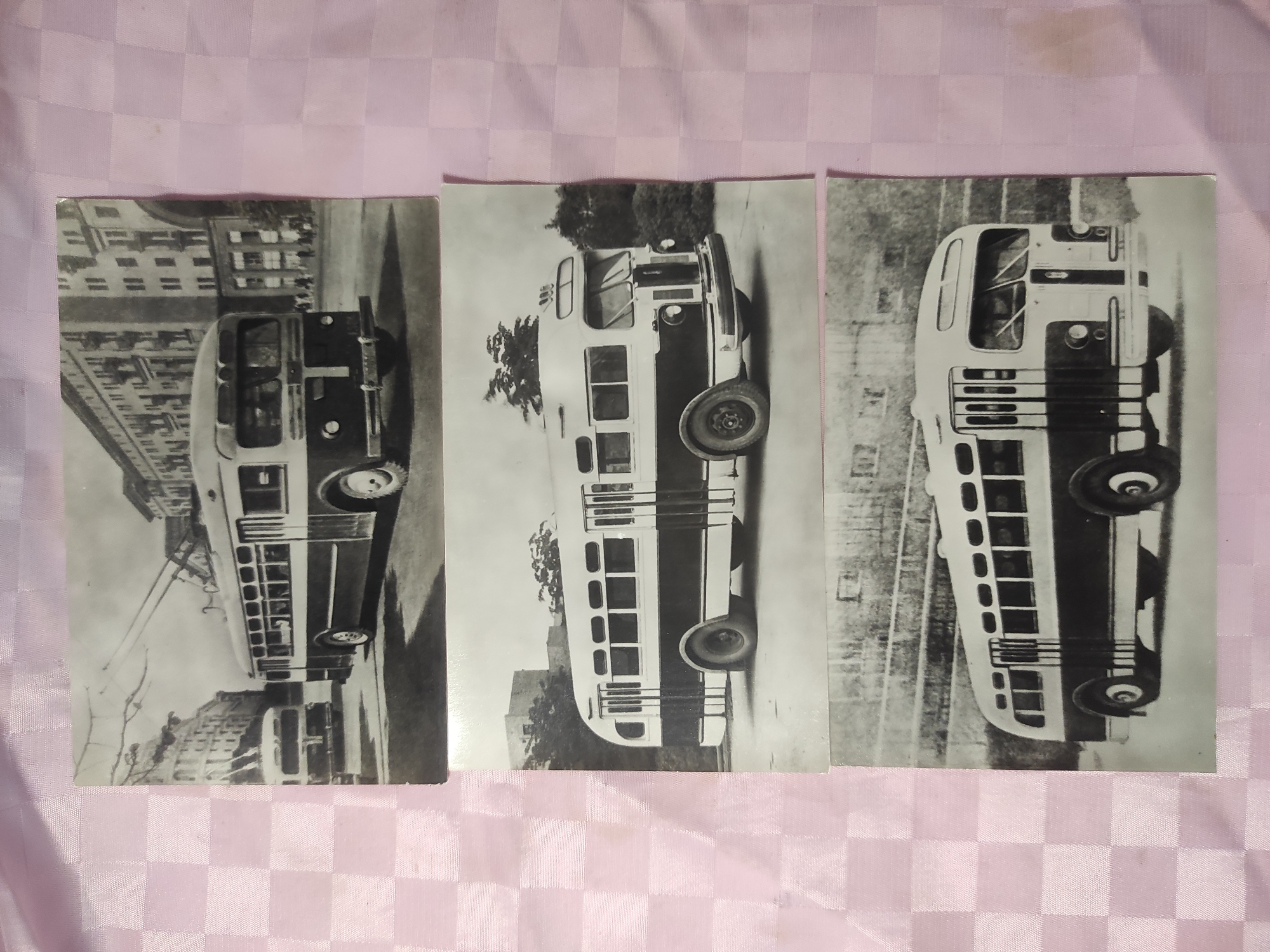 The image displays three black and white photographs of buses, arranged vertically on a pink checkered fabric surface.
The top photograph shows a trolleybus on a city street. The trolleybus is dark colored with a lighter colored roof and trim. It has multiple windows along its side and a rounded front with headlights. Overhead wires for the trolleybus are visible. A second, lighter colored bus is parked in the background to the left. Tall buildings line the street in the background, suggesting an urban environment.
The middle photograph features a bus with a dark lower body and a lighter upper section. It has a series of rectangular windows along its side and a rounded front with large headlights. The bus appears to be parked or moving slowly on a paved surface, with some trees and buildings visible in the background.
The bottom photograph shows a bus that is similar in design to the middle one, with a dark lower body and a lighter upper section, and a similar window configuration. The front of this bus is visible, with a windshield, headlights, and a grille. The bus is on a paved road, with lush greenery and trees in the background.
There is no information within the image itself that would allow for identification of the location as Chernihiv, Ukraine.
