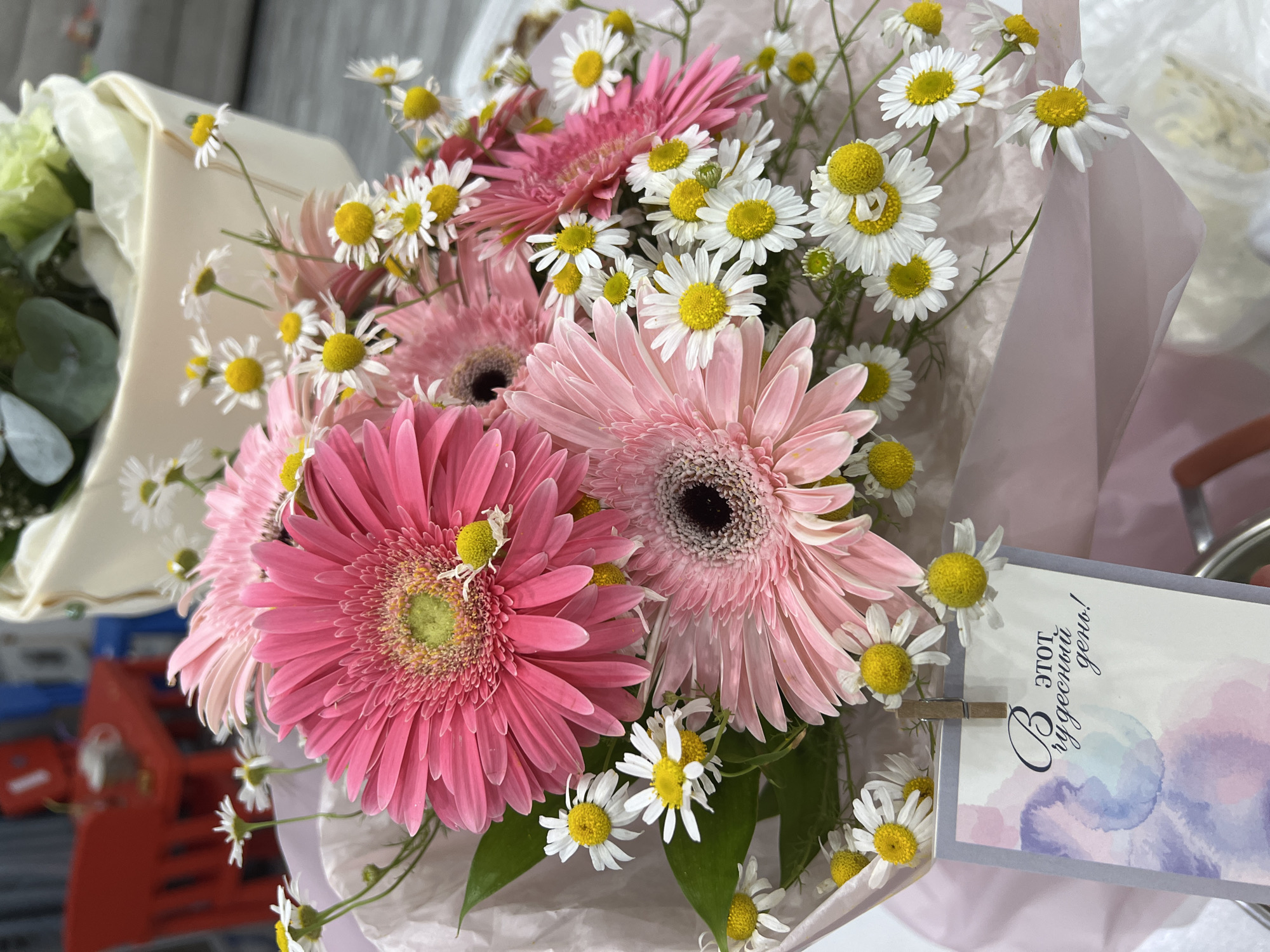 A bouquet of pink gerbera daisies and white daisies is presented, wrapped in light pink paper. A small card attached to the bouquet has the Russian text "В этот чудесный день!" which translates to "On this wonderful day!". The background is blurred but appears to be an indoor setting with hints of blue and red objects, possibly shopping carts, suggesting a retail or market environment in Bataysk, Russia. The lighting suggests daytime.