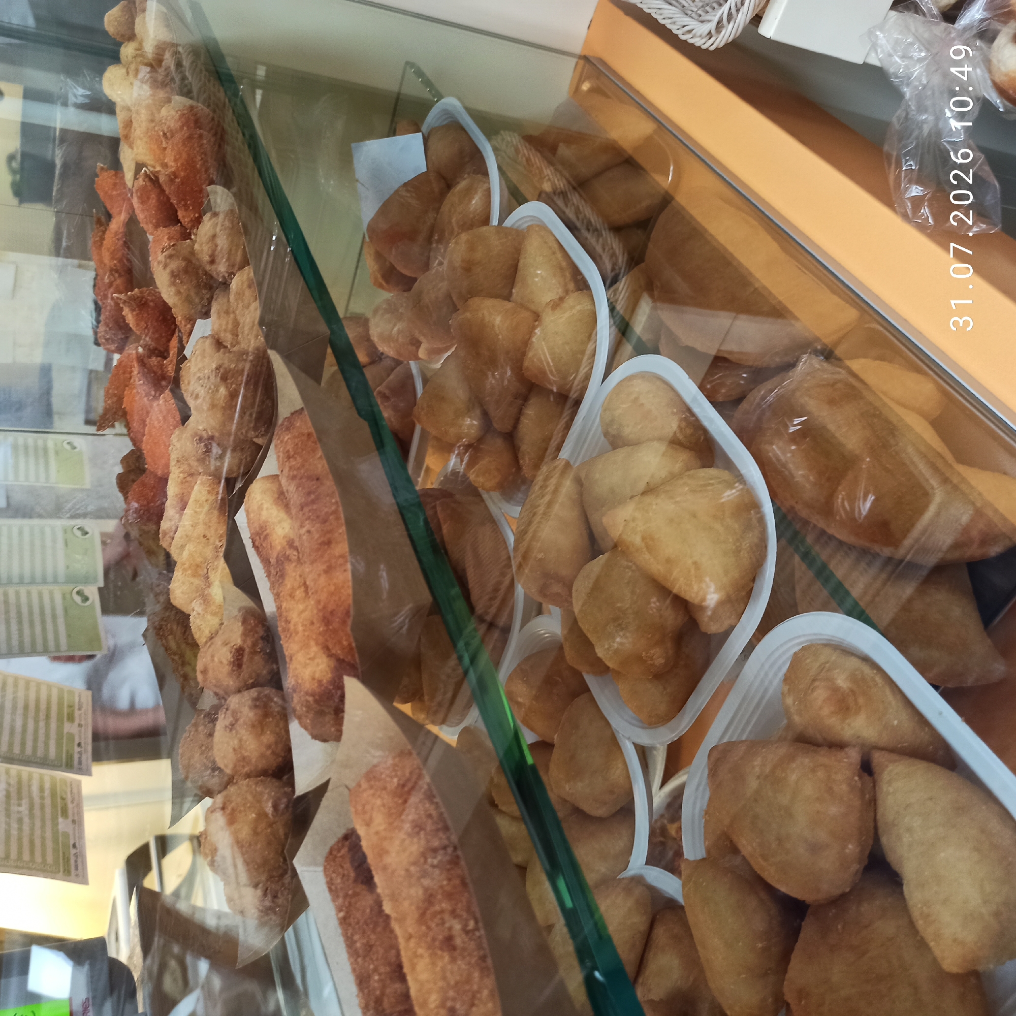 The image shows a display case filled with various fried pastries and breads at a bakery in Modugno, Italy. The pastries are arranged in white plastic trays and paper liners. The display case is made of glass and reflects the interior of the establishment. In the background, there are signs with text and a person partially visible. The timestamp "31.07.2026 10:49" is overlaid on the bottom right corner of the image.
