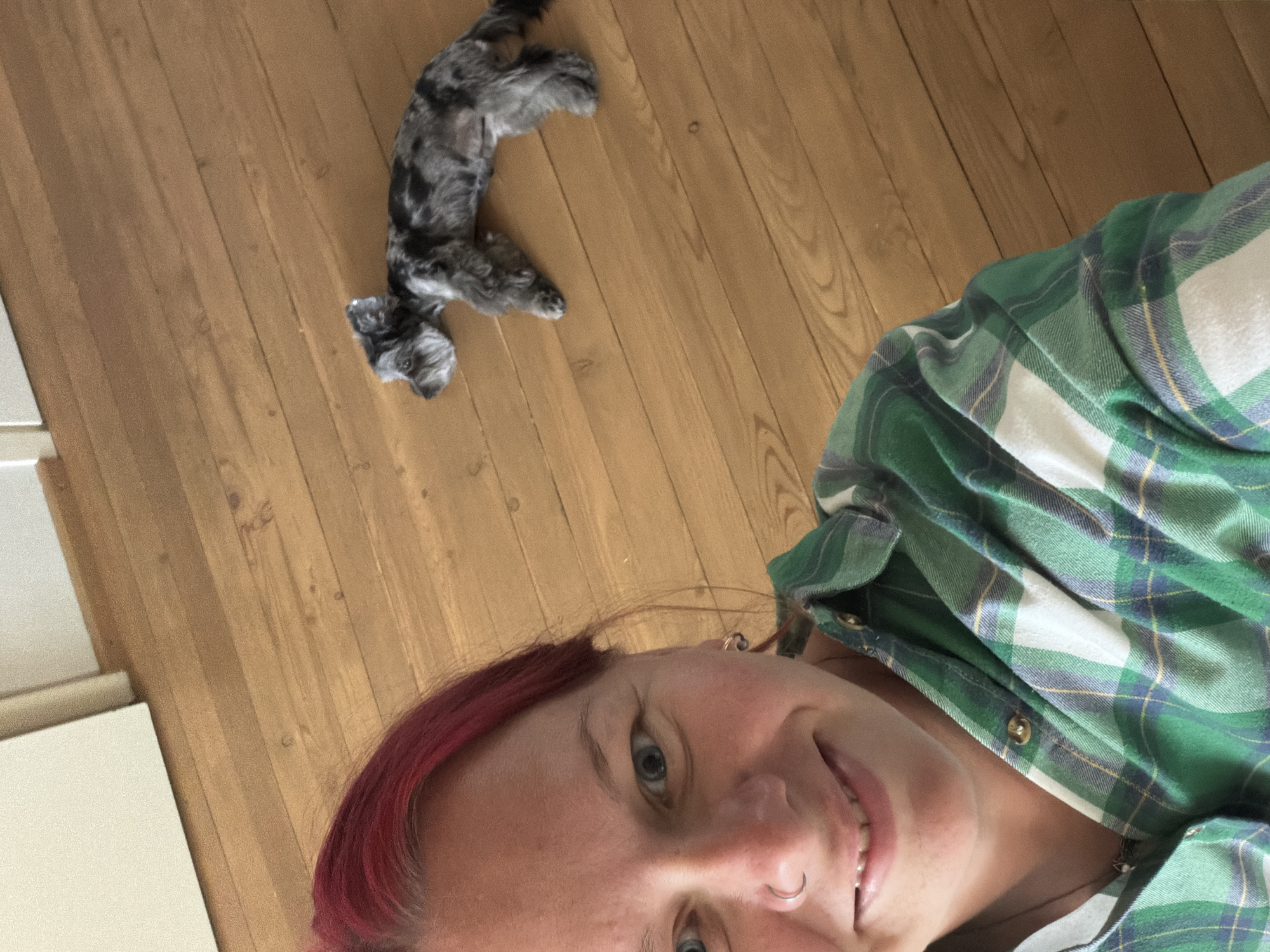 A woman with bright red hair and blue eyes smiles at the camera. She is wearing a green and white plaid shirt. A small dog with grey and black fur lies on a wooden floor to her right. The background consists of wooden floorboards and a light-colored wall. The image appears to be taken indoors, possibly in a home. There is no information to indicate this scene is located in Chaikovsky, Russia.