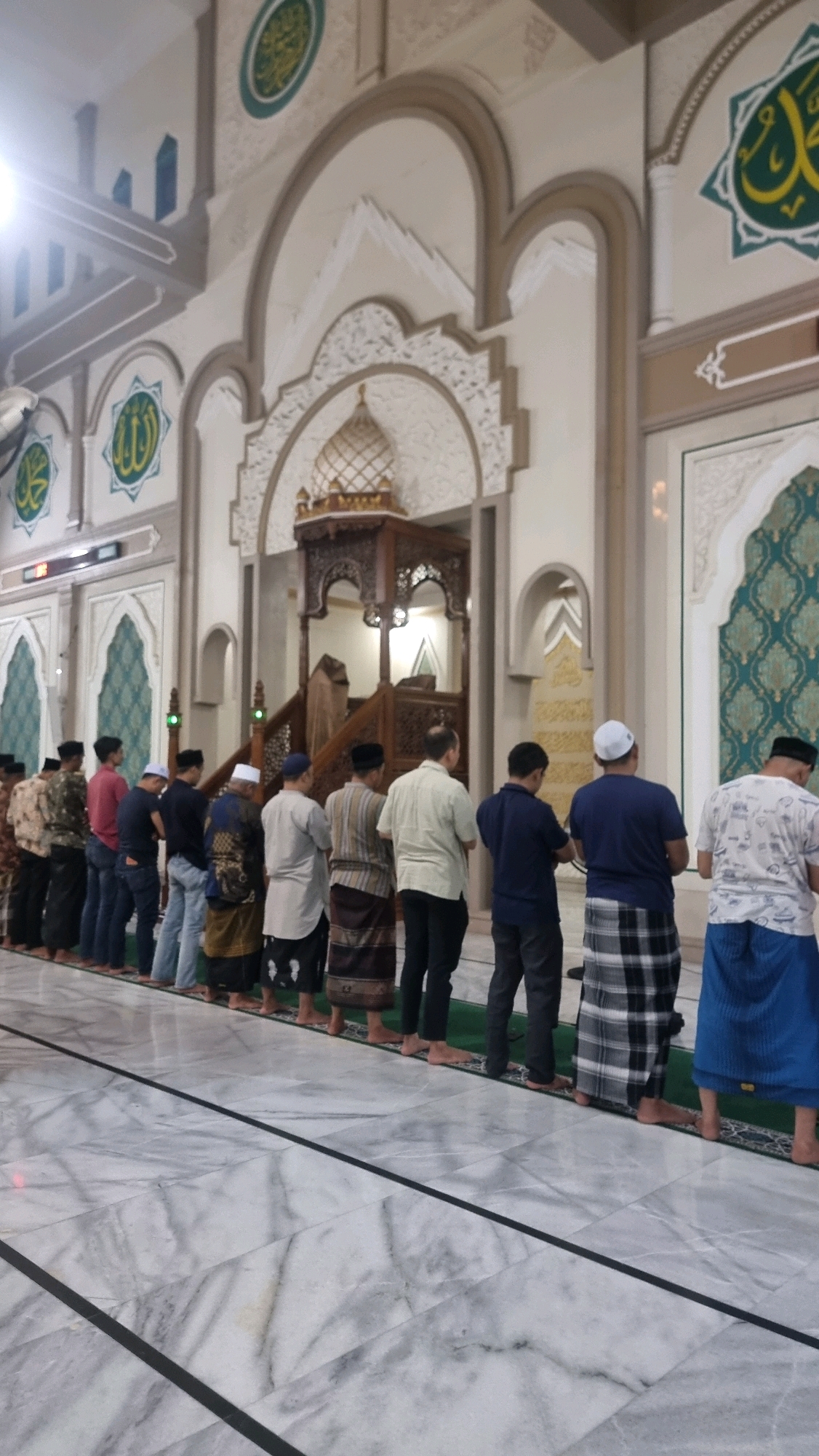A vertical, medium shot captures a group of men performing what appears to be Islamic prayer (Salat) inside a mosque. The men are lined up in rows facing a wooden minbar (pulpit), which is ornately carved and topped with a golden dome. The mosque's interior is decorated with intricate patterns, including Islamic calligraphy and geometric designs in green and gold. The floor is made of polished marble with dark lines delineating sections. The men are dressed in traditional Islamic attire, including sarongs and caps. The lighting is soft, suggesting an indoor environment. The overall impression is one of solemnity and devotion. Based on the context of the image and its presentation, this scene is likely from the Baiturrahman Grand Mosque in Lhokseumawe, Indonesia, a prominent religious and architectural landmark in the city.