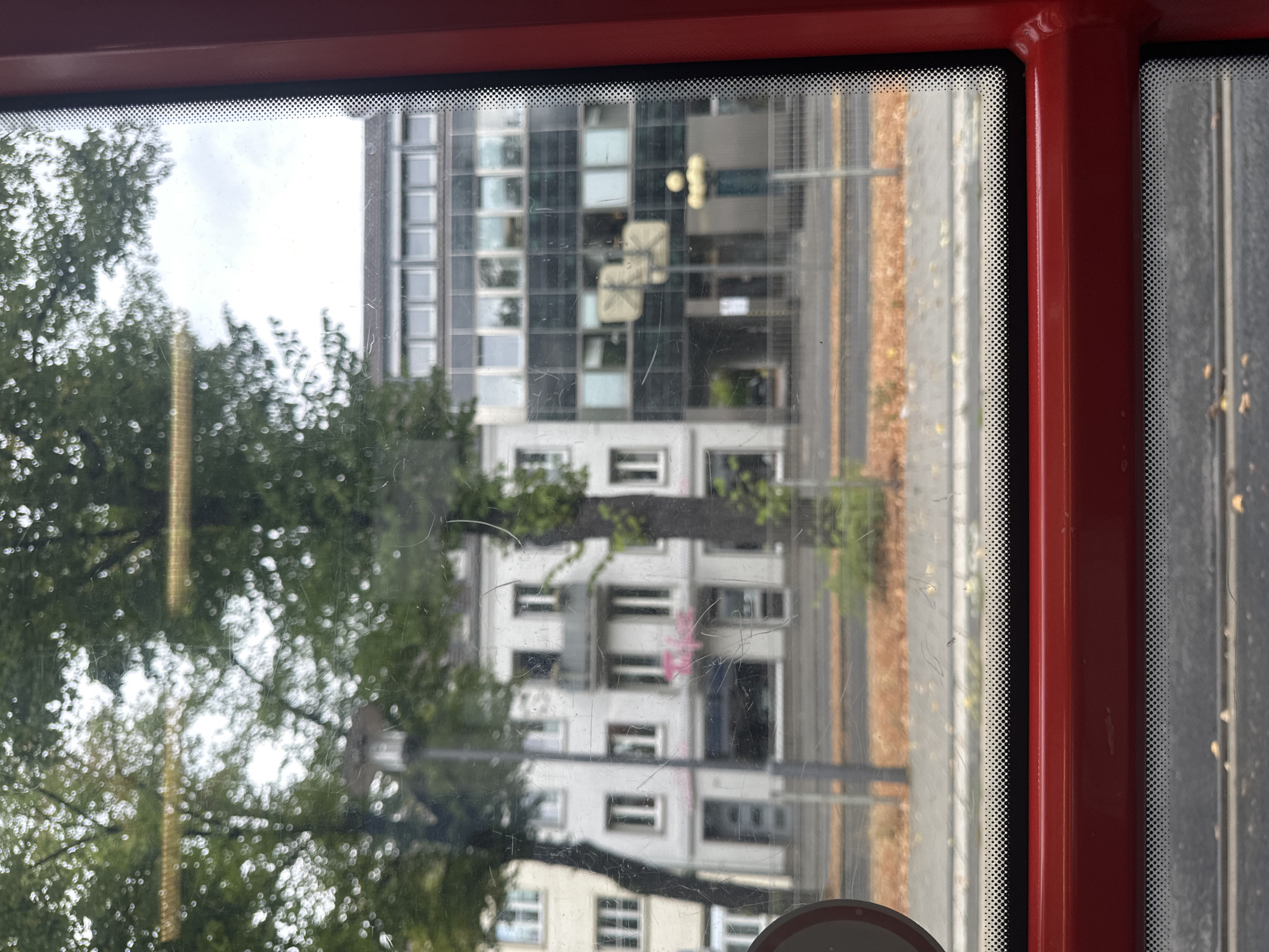 This image was taken in Krefeld, Germany, likely from the window of a vehicle. The foreground shows the red interior of a vehicle, including a red bar and the edge of a window frame. The window itself is dirty and scratched, with reflections of trees and the sky visible. Beyond the window, a street scene unfolds. On the left, there is a light-colored building with several windows and a balcony. To the right of this building is a darker, modern building with multiple floors and numerous windows. Trees line the street, their leaves creating a blurred effect due to the window's condition. The ground appears to be a sidewalk or street with fallen leaves scattered on it. There are no people clearly visible in the image, and the overall impression is one of a typical urban environment.