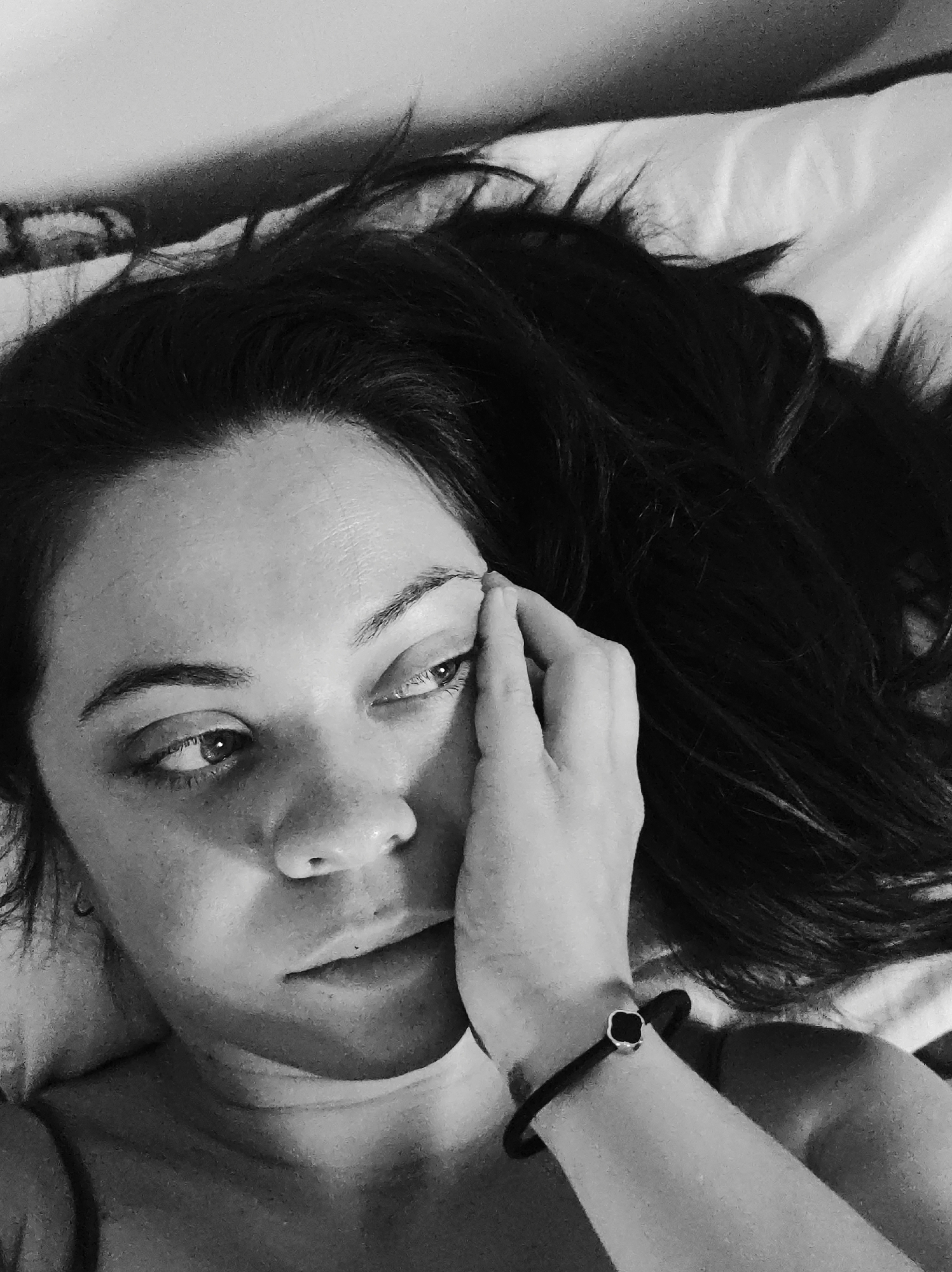 A black and white, close-up, overhead photograph depicts an individual lying down indoors. The subject, with dark hair cascading around their head, is positioned on a light-colored surface, likely bedding or a pillow. Their eyes are open and directed towards the upper left of the frame, with a slightly distant or pensive gaze. The subject's right hand is placed against their right cheek and temple, with fingers extending towards the right eyebrow. A dark bracelet featuring a light-edged, dark square or clover-shaped charm is visible on the subject's left wrist. The left shoulder and a portion of the chest are bare or lightly clothed with a thin strap visible. A faint, darker patterned object, possibly a headboard or wall hanging, is partially visible in the upper left background. The scene is an unidentified indoor setting, with no specific city information discernible.
