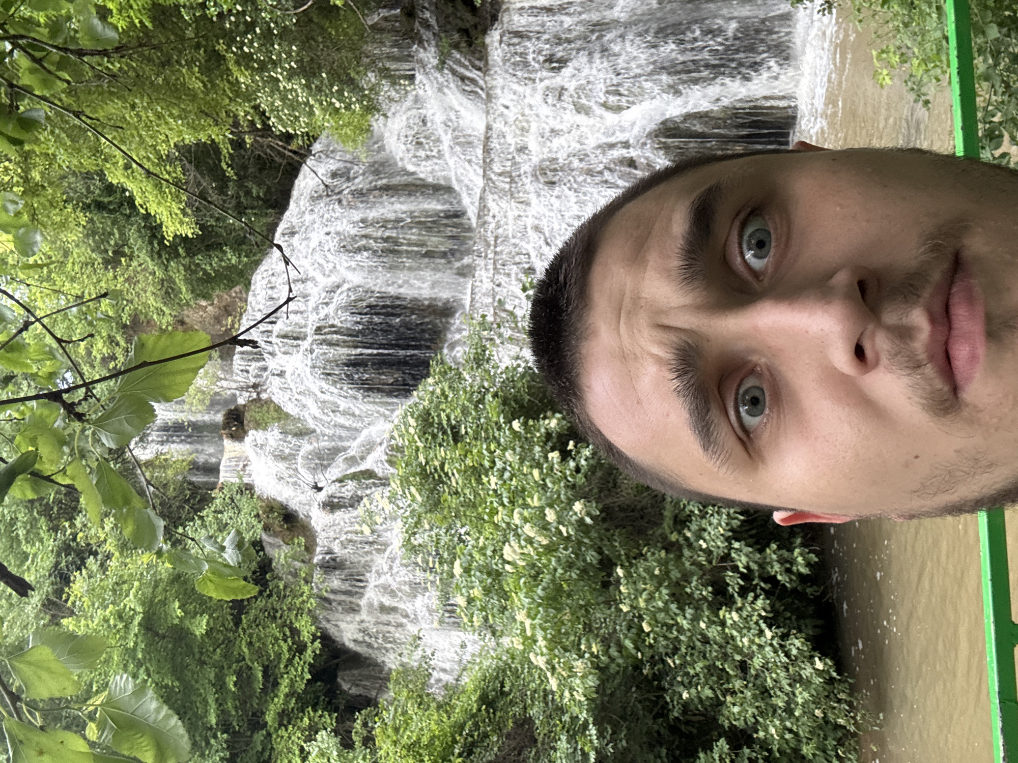 A man is taking a selfie in Krushuna, Bulgaria with the Krushuna Waterfalls in the background. He has light skin, short dark hair, and blue eyes. The waterfalls are cascading down rocks surrounded by green vegetation. A body of water is also visible.
