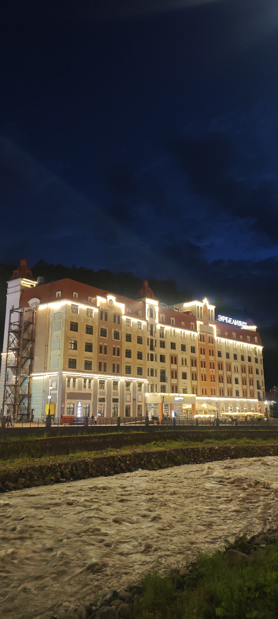 The photograph captures a view of the "ЭРБЕЛИЯ" hotel at night, situated in Estosadok, Russia. The multi-story hotel is illuminated with exterior lighting, highlighting its architectural details and the hotel's name sign on the roof. A rapidly flowing river occupies the foreground, reflecting the lights of the building and the sky, which appears to be a deep blue with hints of clouds. A low barrier separates the riverbank from a narrow strip of grass and a walkway in front of the hotel. People are visible along the walkway, suggesting activity in the area. Scaffolding or a fire escape is attached to the leftmost section of the building. The overall environment suggests a calm yet dynamic evening scene.