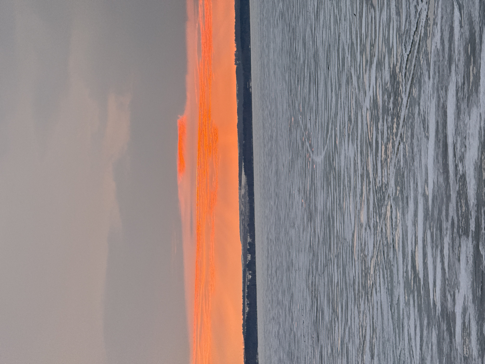 The image depicts a scene at sunset in Votkinsk, Russia, showcasing a partially frozen body of water, likely a lake or river. The ice surface exhibits numerous skate tracks and patches of snow. A dark, tree-lined horizon separates the icy expanse from the sky, which displays a dramatic gradient of grey, orange, and yellow hues, indicative of the setting sun. The scene is devoid of people.