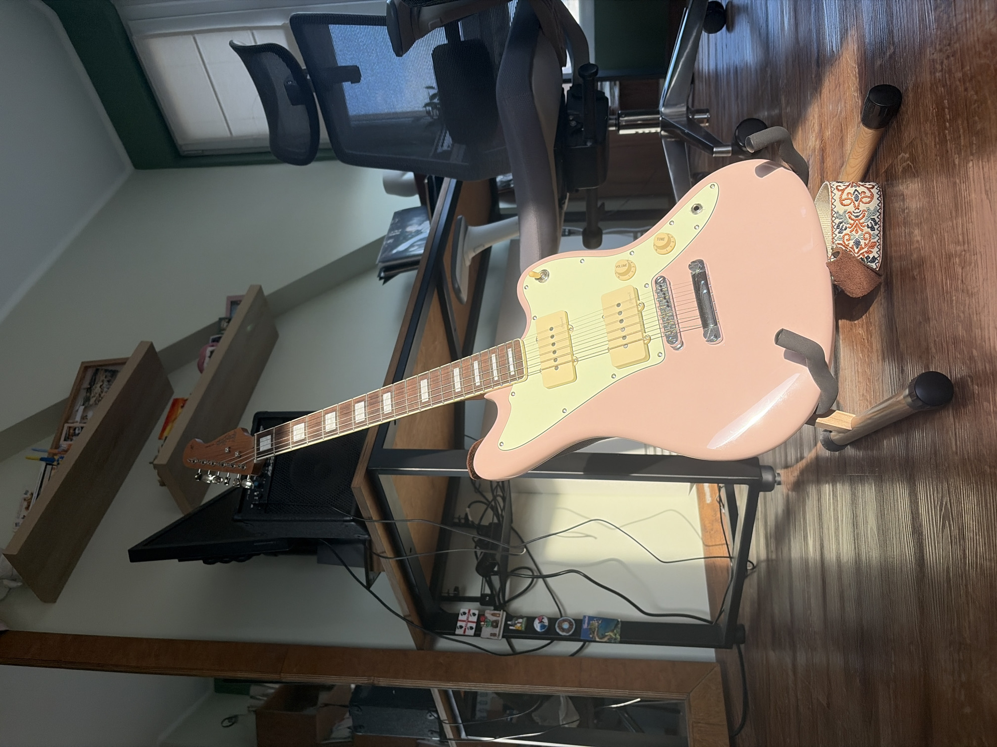 The image depicts an interior room, specifically a home office setup, situated in Unknown, Unknown. In the foreground, a glossy pink electric guitar with a light yellow pickguard, two cream-colored humbucker pickups, and matching control knobs rests on a wooden guitar stand with black rubberized supports. A decorative fabric guitar strap, patterned with orange, blue, and brown motifs, is positioned at the base of the guitar stand. The floor is composed of dark brown wooden planks, illuminated by natural sunlight entering from the right, casting distinct shadows.
In the midground, a desk with a black metal frame and a light wooden surface is visible. On the desk, a black computer monitor is positioned to the left of a black guitar amplifier. Below the desk, a tangle of black electrical cables is visible. Attached to a vertical surface under the desk are several decorative magnets, including one depicting the flag of Georgia (red cross on white field) and another featuring a green island. To the far left, a tall, dark wooden-framed mirror or panel is partially visible.
The background features a light green wall adorned with two light wood floating shelves, on which small items such as framed photographs and a white figurine are displayed. To the right, a black mesh office chair with a metallic five-star base and wheels is positioned in front of a window equipped with white horizontal blinds. A dark green molding runs along the top of the window frame. No human subjects are present in the image.