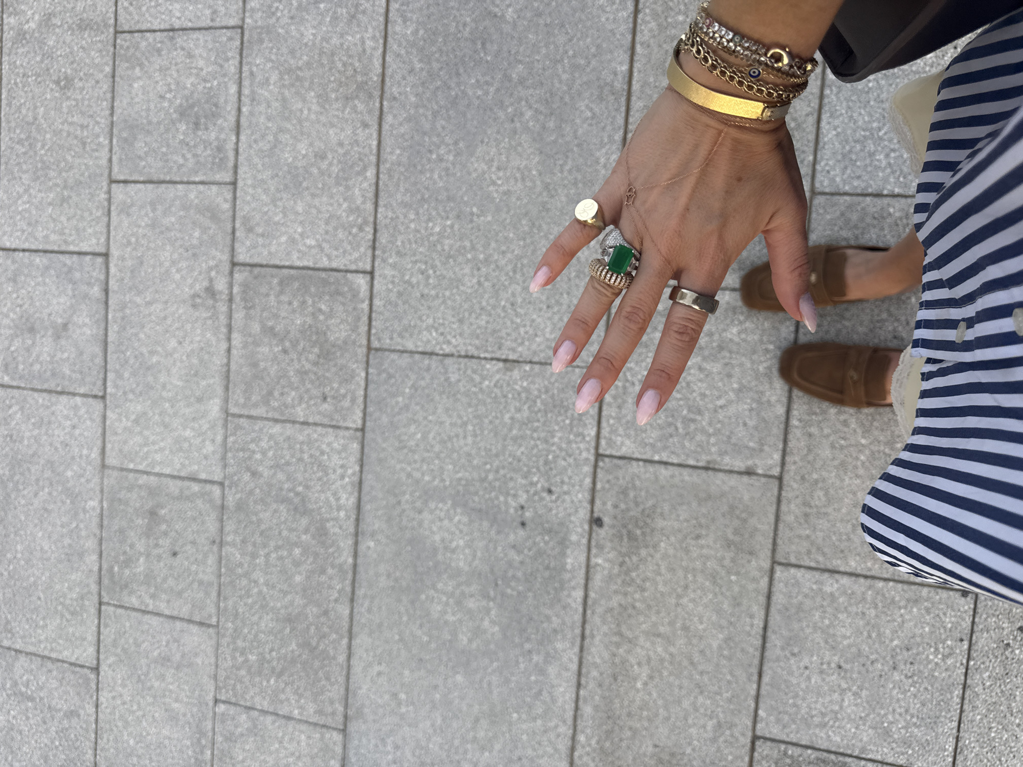 The image shows a person standing on a paved surface, likely a pedestrian walkway. The paving consists of large, rectangular, light grey stones arranged in a running bond pattern. The person's hand is prominently displayed, adorned with several gold bracelets and rings, including one with a large emerald-cut green stone. A portion of the person's outfit is visible, including a striped blue and white garment and brown leather loafers. The style of paving, the fashion items, and the overall aesthetic suggest an urban environment, possibly in a high-end area. There is no explicit indication of Dubai, United Arab Emirates, in the image.