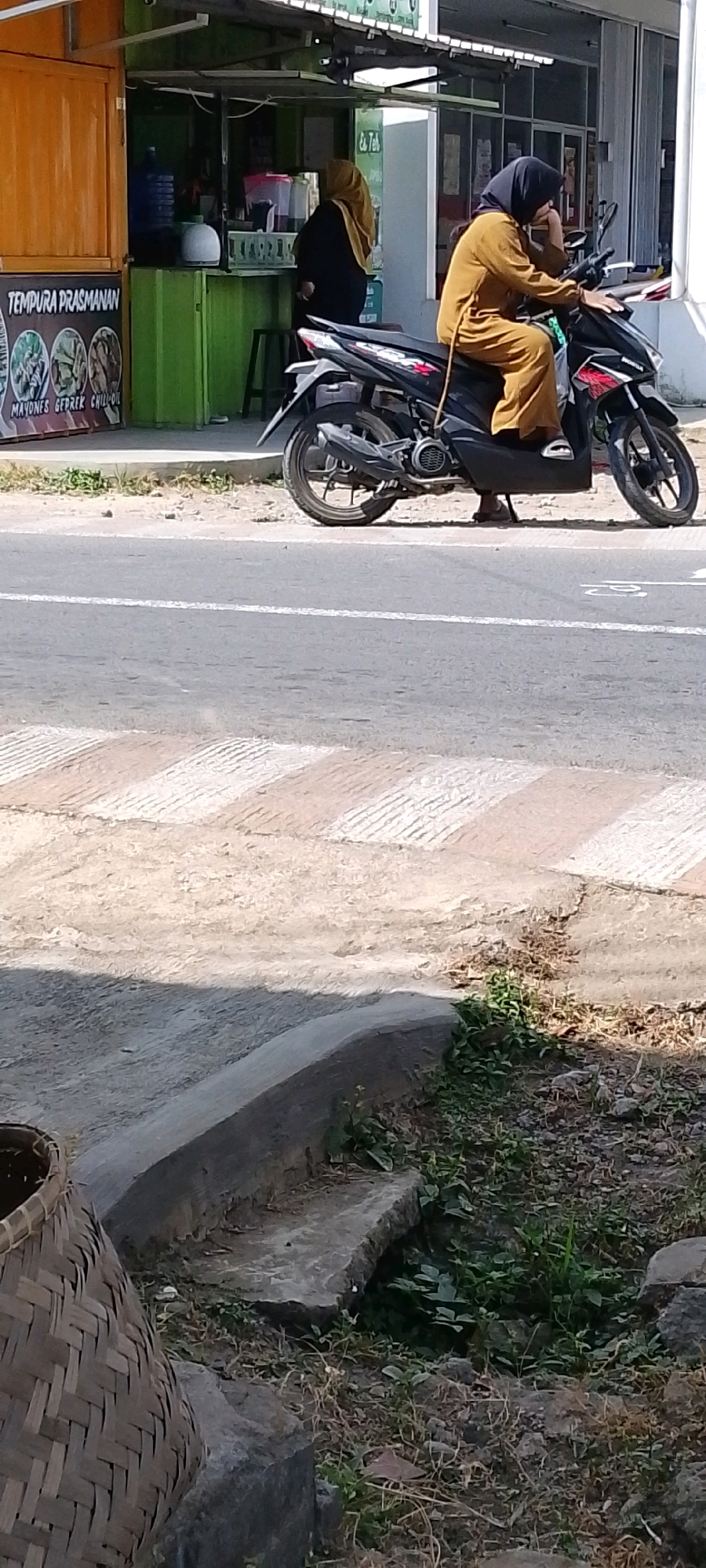 The image depicts a street scene in Kertahayu, Indonesia. A woman wearing a brown hijab and a matching outfit is seated on a black scooter. She is holding her hand to her face, appearing to be in conversation or deep thought. The scooter is parked on the side of a road with a pedestrian crossing marked by white and brown stripes. Behind the scooter, a food stall is visible, with a green structure and a sign displaying "TEMPURA PRASMANAN" along with images of food. Another woman, wearing a yellow hijab and a dark outfit, stands near the food stall. The environment appears to be outdoors, with evidence of greenery and dry foliage on the ground. A woven basket is partially visible in the lower left corner of the image. The overall impression is a snapshot of daily life in a local market or street-side vending area.