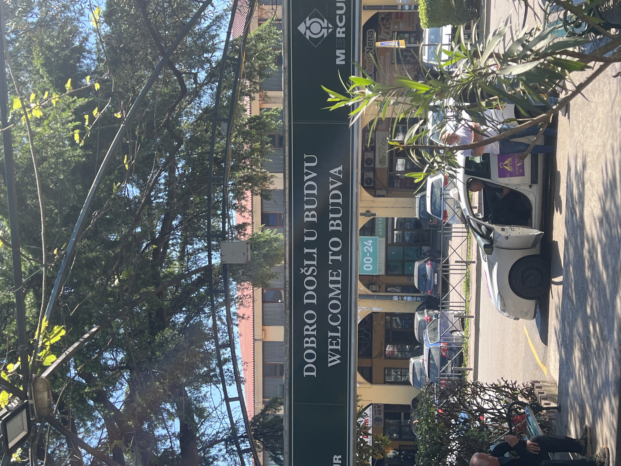 This image captures a vibrant, sunny street scene in Budva, Montenegro, dominated by a prominent welcome sign. The large green sign centrally located in the upper half of the image proudly displays "DOBRO DOŠLI U BUDVU" and "WELCOME TO BUDVA," topped by a "MERCURI" logo.
The scene unfolds on a bright day, indicated by strong shadows on the pavement and clear blue skies visible through dense tree foliage on the left. On the right side of the street, several cars are parked, including a white car with its front door ajar, near which a man in a white t-shirt and jeans stands. A purple sticker on the car suggests it might be a taxi or ride-share service. In the immediate foreground, another man is seated on a bench, facing away from the camera.
Behind the parked vehicles, a row of commercial buildings houses various establishments. Visible signs include "00-24" (likely operating hours), "VEGAPHARM APOTE" (a pharmacy), "kafe. priča" (a cafe), and "kod Nena" with "sarajevska uregdžinica" (a deli or eatery). A sign for "MOKER RENT A CAR" is also visible. The overall impression is a lively urban environment, typical of a tourist town.