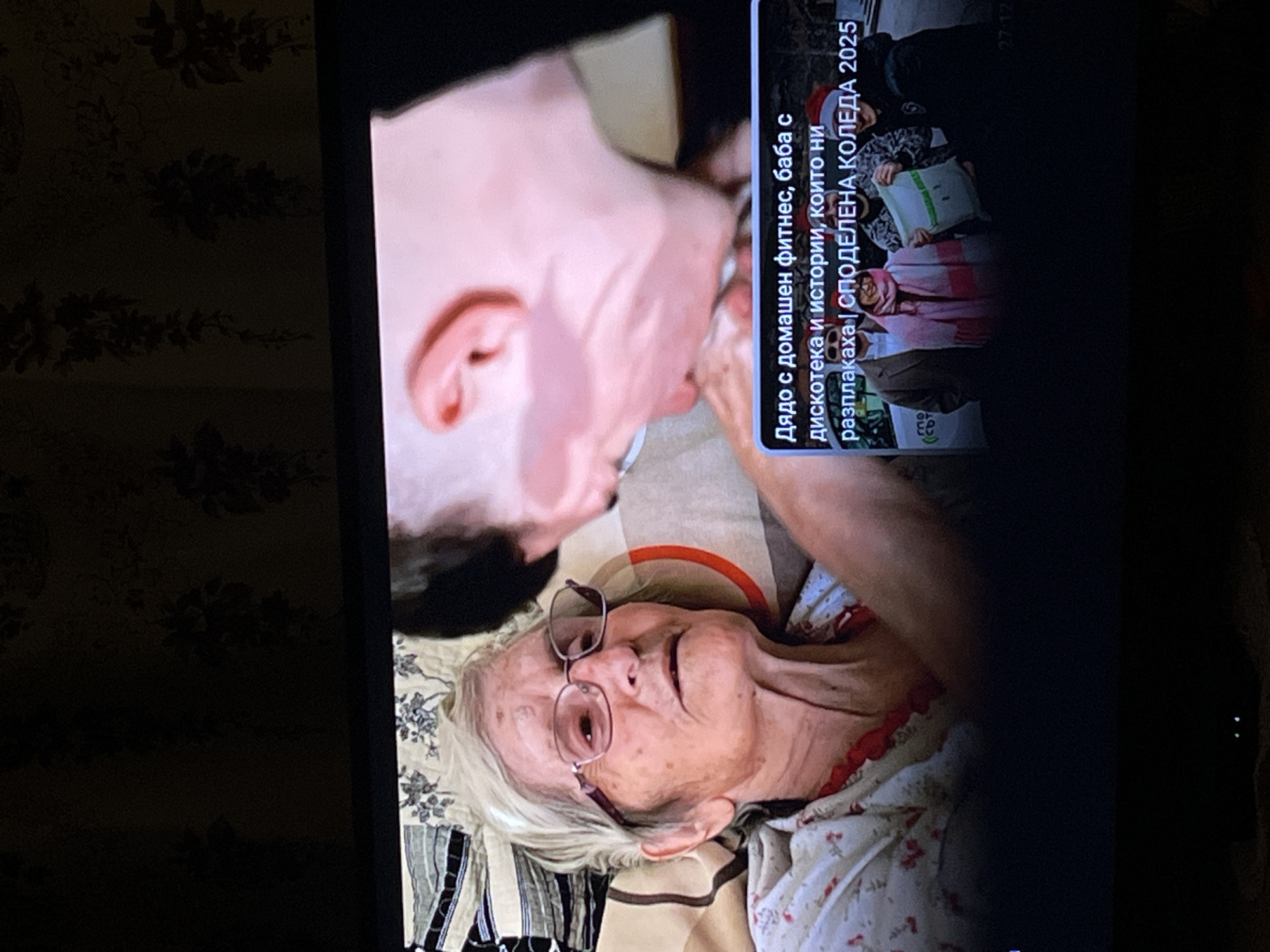 The image captures a screen displaying a scene with an elderly woman wearing glasses, lying down on a patterned surface, possibly a bed. Above her is the face of a younger person. On the right side of the screen, text in Bulgarian reads: "Дядо с домашен фитнес, баба с дискотека и истории, които ни разплакаха | СПОДЕЛЕНА КОЛЕДА 2025" which translates to "Grandpa with home fitness, grandma with disco and stories that made us cry | SHARED CHRISTMAS 2025". The number "27-12" is also visible. The scene is presented on a television or computer screen. The location of Lozarevo, Bulgaria, is not visually identifiable in the image.