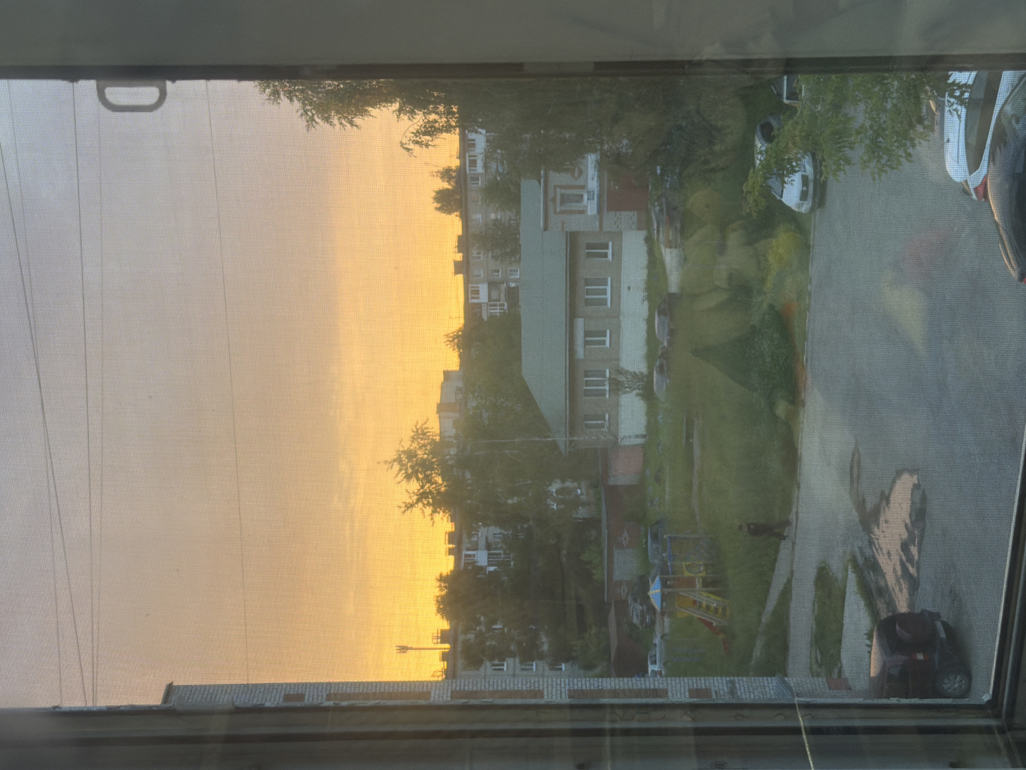 This is a photograph taken through a window screen, likely at sunset, given the warm, golden light illuminating the sky. The image depicts a residential area, possibly an apartment complex, with multi-story buildings visible among mature trees. The architecture suggests a Soviet-era design.
In the foreground, the texture of the window screen is apparent, creating a slightly blurred effect on the scene beyond. A metal handle, likely part of the window mechanism, is visible in the upper left corner. Below the screen, the edge of a brick windowsill or building is present.
The background reveals a street or courtyard with paved surfaces and grassy areas. Several cars are parked, including a dark-colored SUV or minivan with its rear end prominently visible on the right side of the image. A children's playground with colorful structures, including a slide, is also present. The overall impression is one of a typical urban or suburban residential setting in the late afternoon or early evening. There are no discernible people or text within the frame. The scene conveys a sense of calm and everyday life. The lighting and the muted colors suggest a tranquil atmosphere. The view is oriented vertically, as if the camera was held sideways.