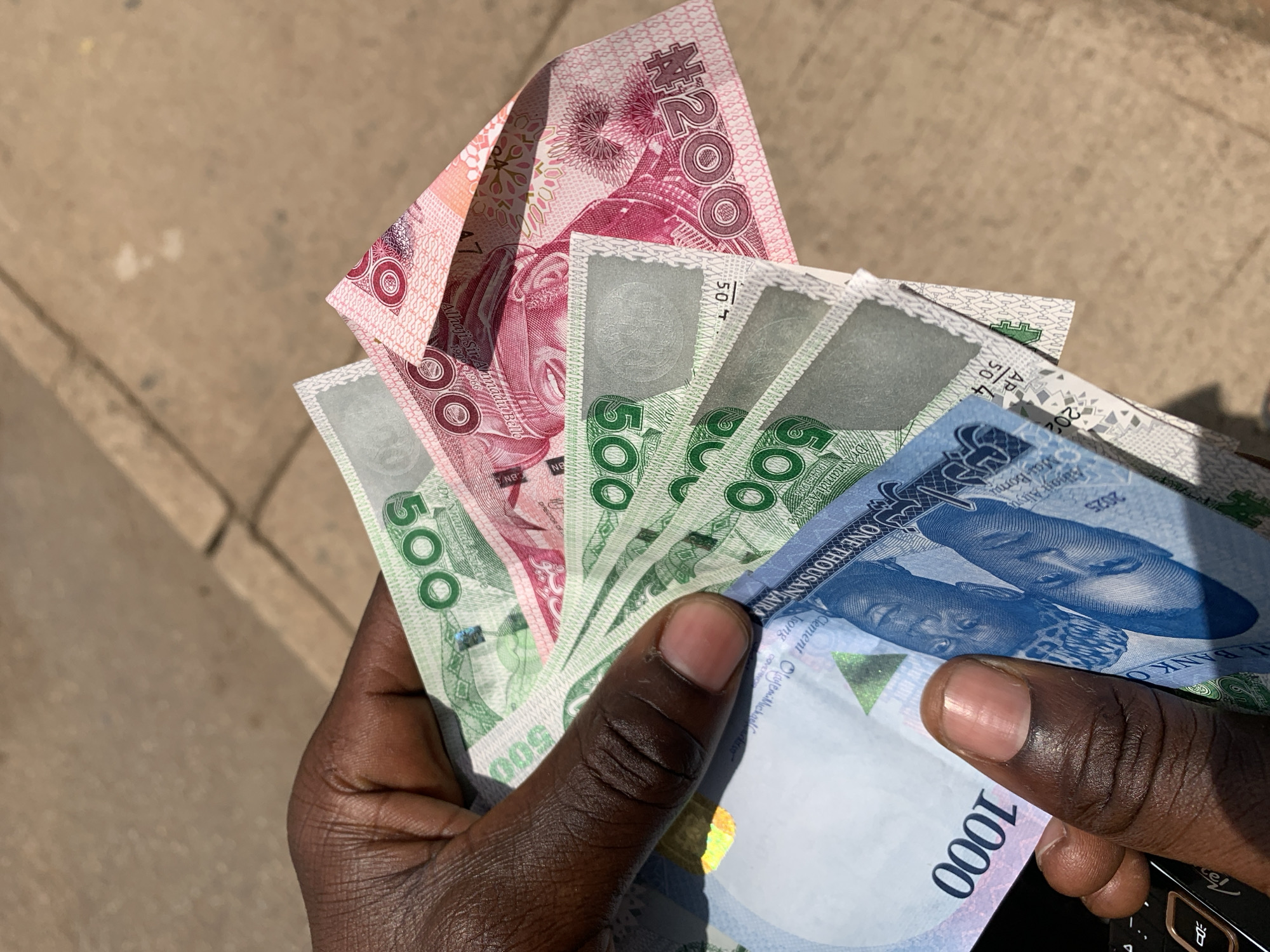 A dark-skinned hand holds Nigerian Naira banknotes. Visible denominations include one ₦200 note, four ₦500 notes, and one ₦1000 note. The photo was apparently taken outdoors on a concrete surface, presumably somewhere in the city of Rafin Par, Nigeria. A portion of a black device is also visible in the lower right corner of the image.