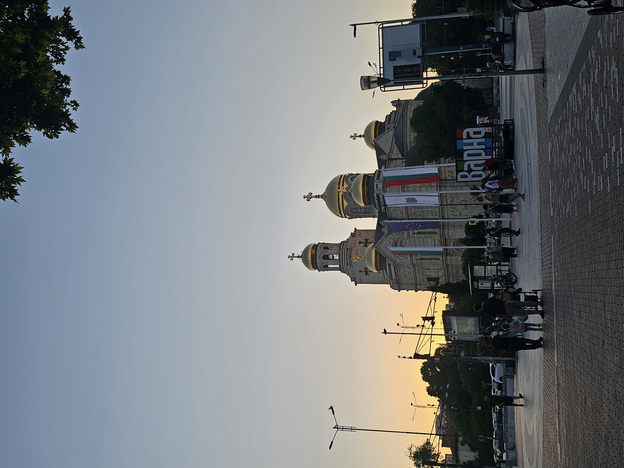 An outdoor, eye-level, vertical shot captures a wide view of the Cathedral of the Assumption of the Virgin in Varna, Bulgaria, during what appears to be twilight. The large, beige stone cathedral dominates the center of the frame, its multiple golden domes and crosses catching the fading light. A tall bell tower with arched openings stands prominently to the left. Three large flags, including the Bulgarian flag, hang from the cathedral's facade. In front of the cathedral, a colorful sign with large, block letters spells out "Варна" (Varna). Several people are scattered throughout the plaza in front of the cathedral, some walking, some gathered in small groups. A family with a stroller is visible in the foreground, with a man and a child standing next to it. To the left of the cathedral, streetlights and traffic signals are present, along with parked cars and trees. The sky is a soft gradient of light blue and orange from the setting sun. The ground is paved with cobblestones in the foreground.