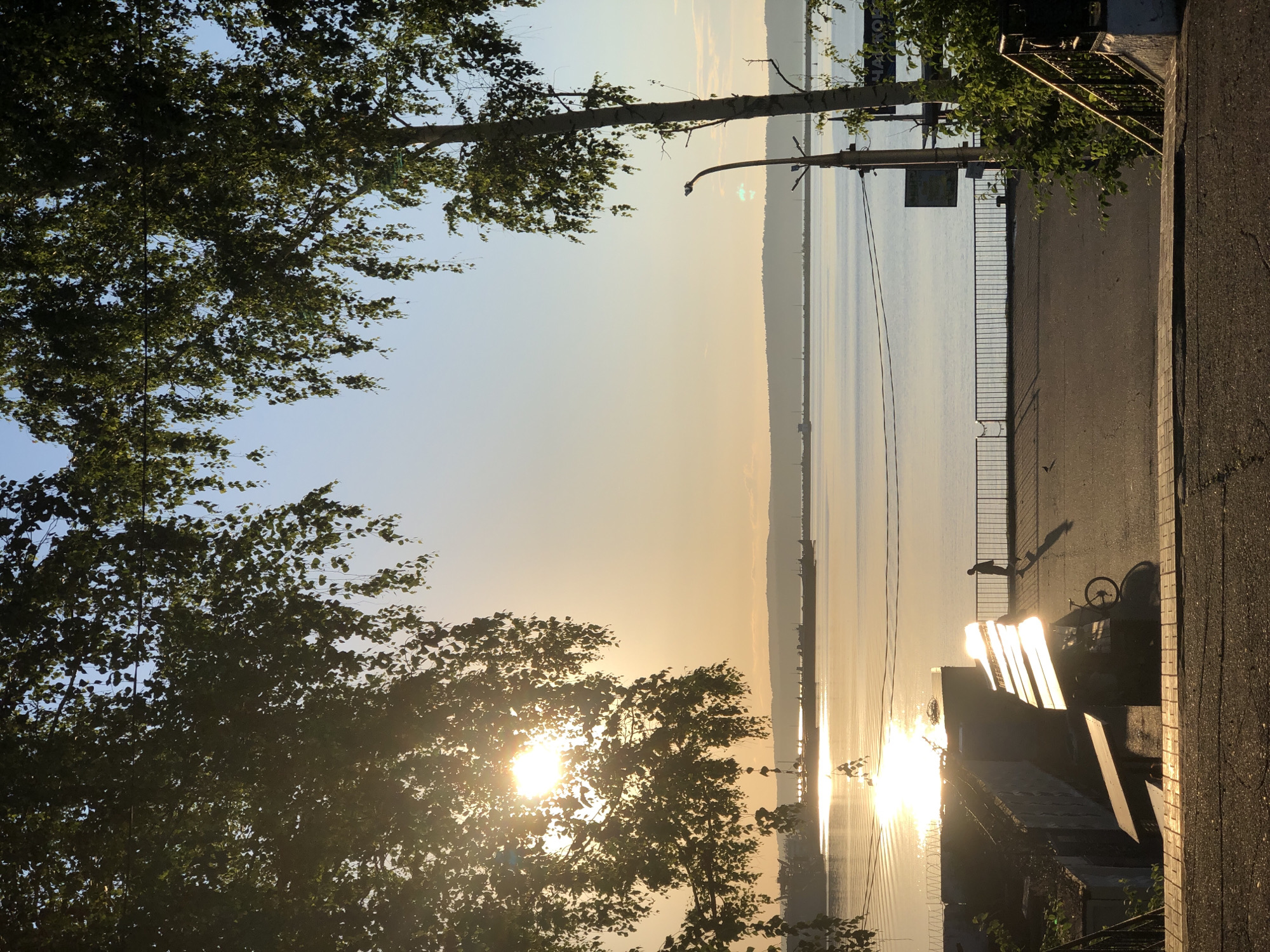 The image depicts a serene sunset scene along the Kama River in Chaikovsky, Russia. The sun, low on the horizon, casts a golden glow across the water, creating shimmering reflections. A lone figure walks along the embankment, silhouetted against the bright light. A bicycle is parked near a bench, suggesting a moment of rest or contemplation. The foreground shows a pathway with steps leading down, while the background is dominated by the vast river and a distant, hazy shoreline dotted with what appear to be industrial structures or ships. Overhanging trees frame the scene, their leaves dappled with sunlight. The sky transitions from a pale blue near the horizon to a deeper hue overhead. The overall atmosphere is peaceful and tranquil, capturing the beauty of a summer evening in Chaikovsky.