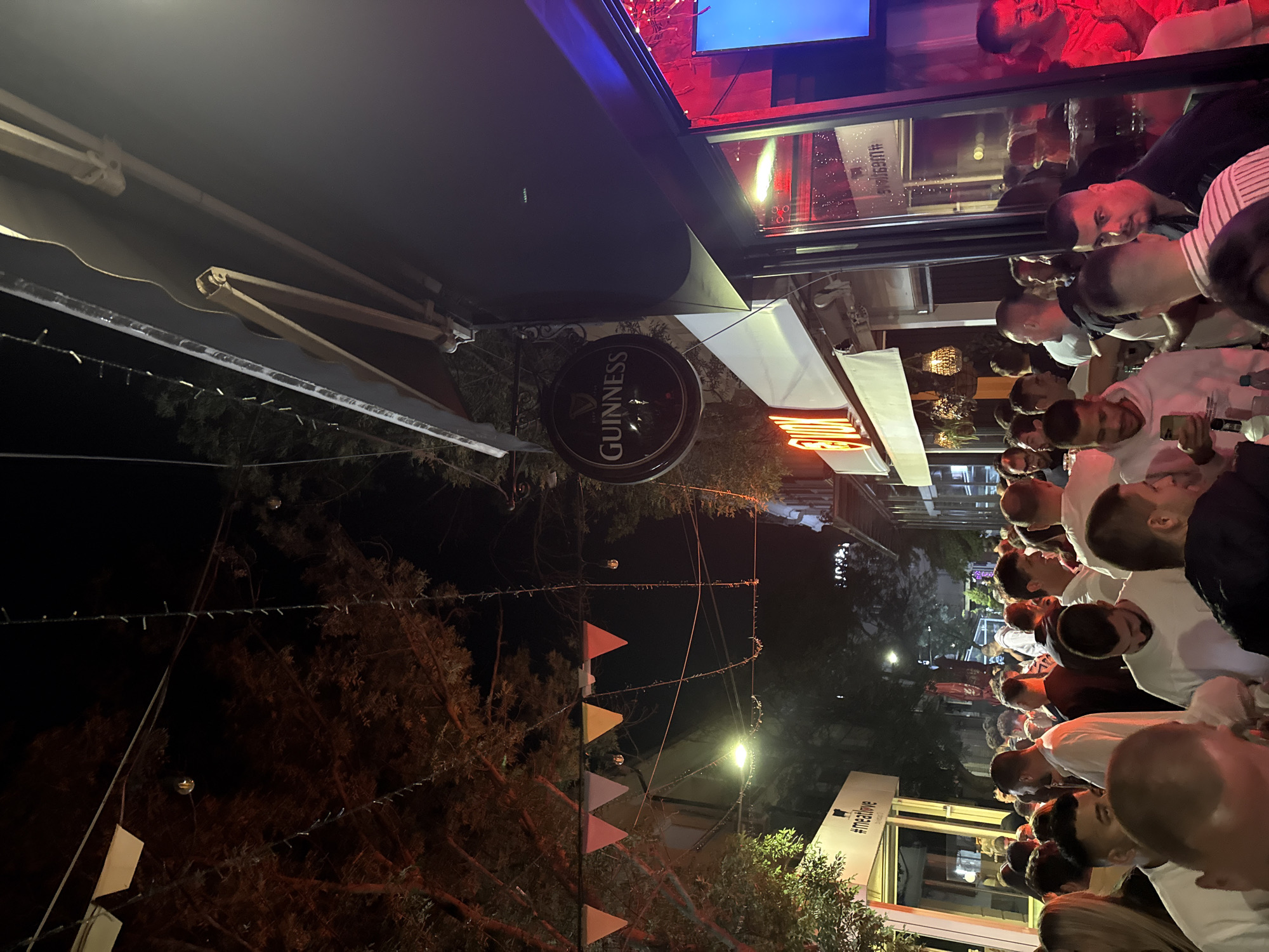 The image depicts a nighttime scene outside a bar or club in Blagoevgrad, Bulgaria. A crowd of people, predominantly young adults, are gathered on a street, with many appearing to be socializing and enjoying themselves. The establishment has a Guinness sign hanging outside, suggesting it is a pub or bar. String lights and triangular pennant banners are strung across the street, adding to the festive atmosphere. The scene appears to be lively and busy, characteristic of a popular nightlife spot. The surrounding buildings and streetlights indicate an urban setting.