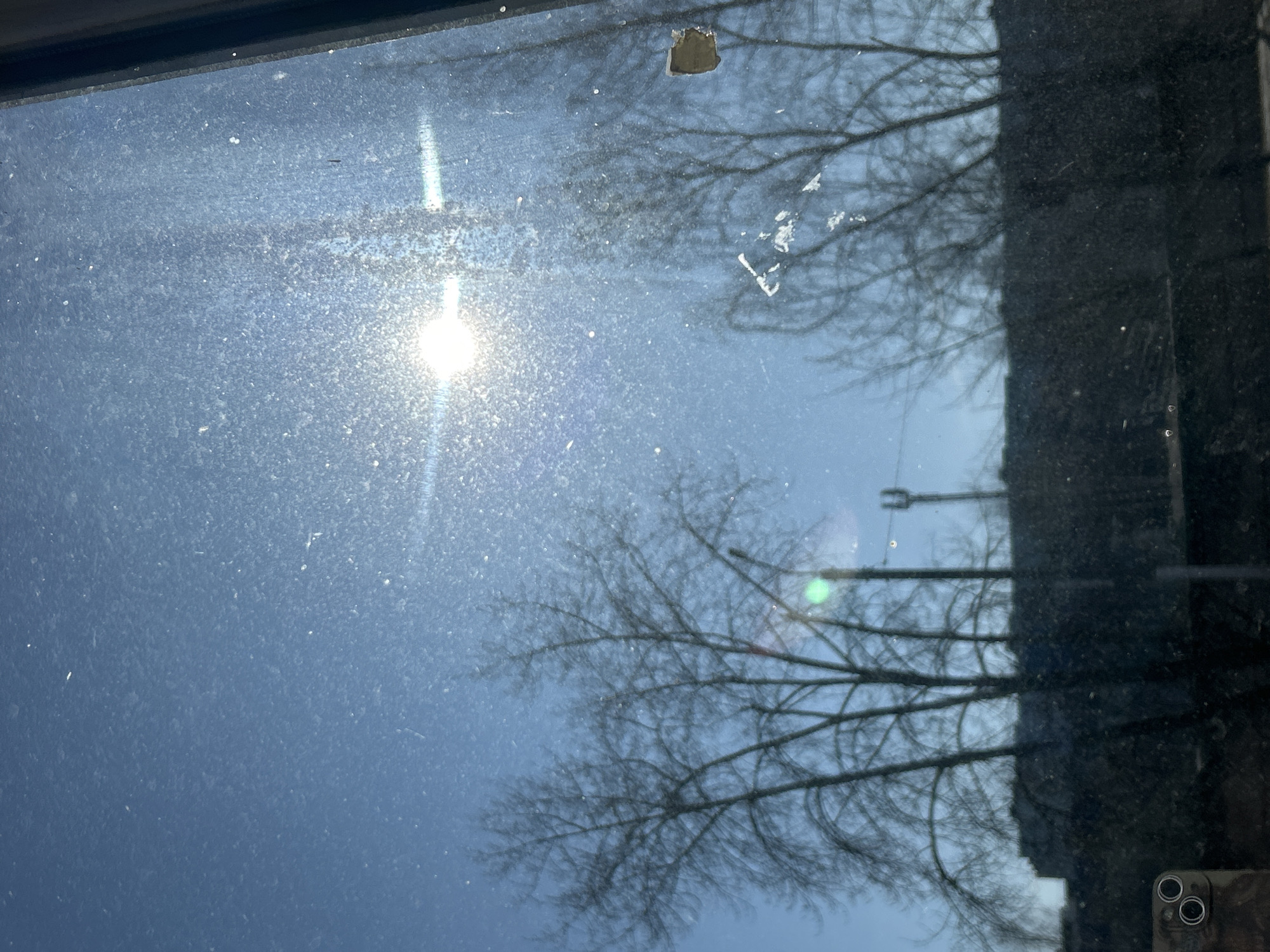 The image shows a view through a dirty window. The sun is visible, creating a glare. Bare tree branches are reflected in the glass. A building is partially visible on the right side of the frame. There is a piece of debris stuck to the window. A power pole is reflected in the glass. The lower right corner shows the camera of a smartphone. It is daytime. The city cannot be determined from the provided information.