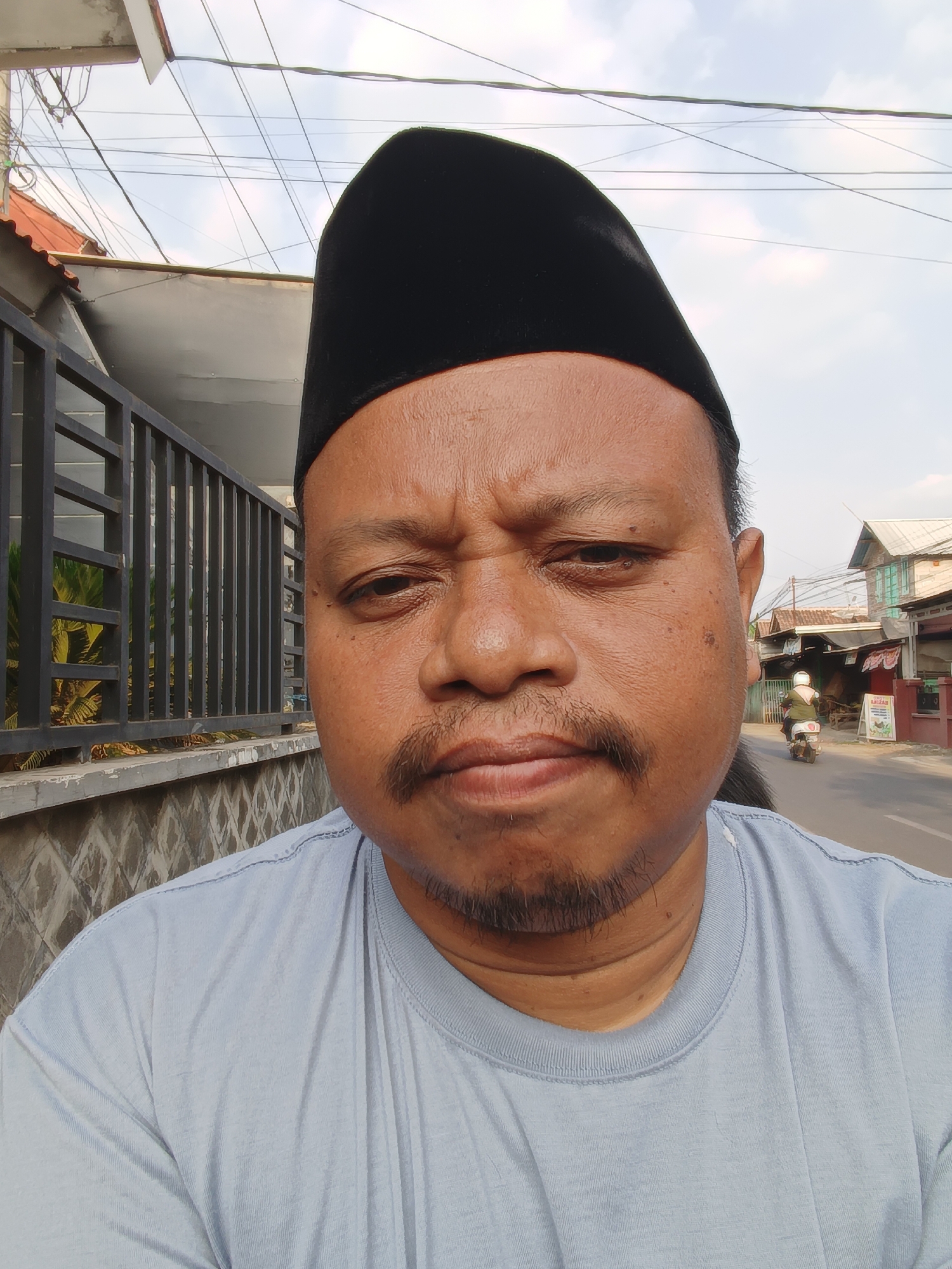 A close-up, selfie-style shot shows a man wearing a black peci hat and a light blue t-shirt. He has a mustache and a goatee. The background suggests an outdoor, urban environment, likely in Kandangjati Wetan, Indonesia, given the presence of a street with traffic and buildings. The man is looking directly at the camera with a somewhat serious expression. The image is taken from the chest up, focusing on the man's face and shoulders. The setting includes a fence and some vegetation on the left, and a street with a person on a motorbike in the distance on the right. Overhead wires are visible against a cloudy sky.