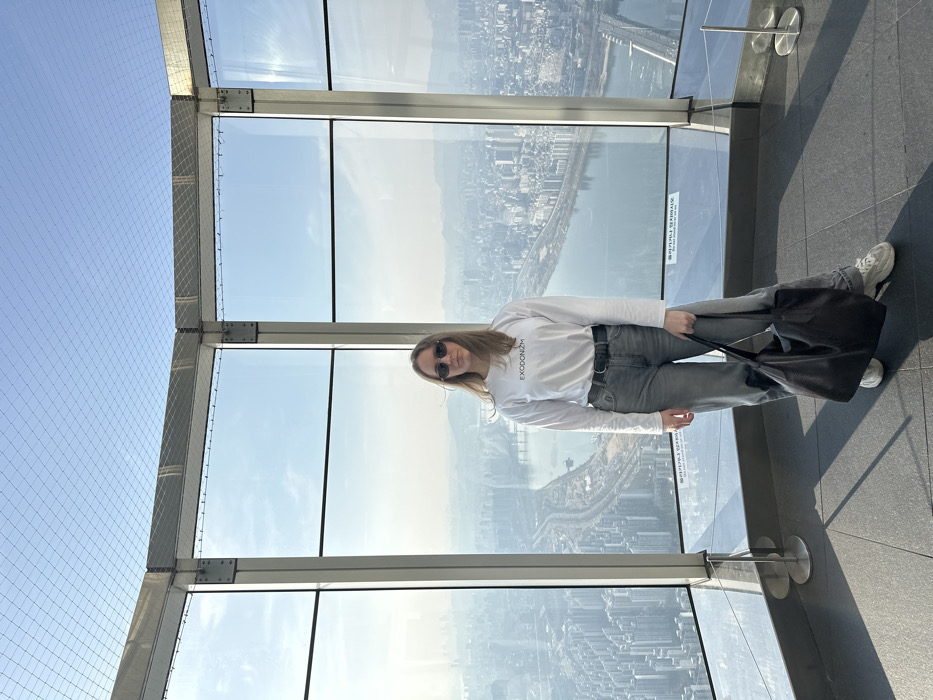 An adult female subject stands centrally within an observation deck located at the Seoul Sky, Lotte World Tower, in Seoul, South Korea. She has light brown hair and wears dark, oval-shaped sunglasses. Her clothing consists of a white long-sleeved shirt with the word "EXODONISM" printed in black on the chest, light grey denim jeans secured by a dark belt, and white athletic shoes. She holds a large, black tote bag in her left hand.
The observation deck features a faceted wall of large, clear glass window panels, framed by dark grey or black metal supports that extend vertically and horizontally. A metal grid structure and safety netting are visible above the main window sections. The floor is a dark grey, possibly tiled, surface. Beyond the glass, a panoramic view of the sprawling city of Seoul unfolds under a clear blue sky with a slight haze on the horizon. The Han River is prominently visible, winding through the urban landscape, flanked by dense clusters of high-rise residential and commercial buildings. Multiple bridges span the river. Sunlight illuminates the scene, casting subtle shadows on the observation deck floor. Small, cylindrical metallic floor markers are present near the base of the window frames.