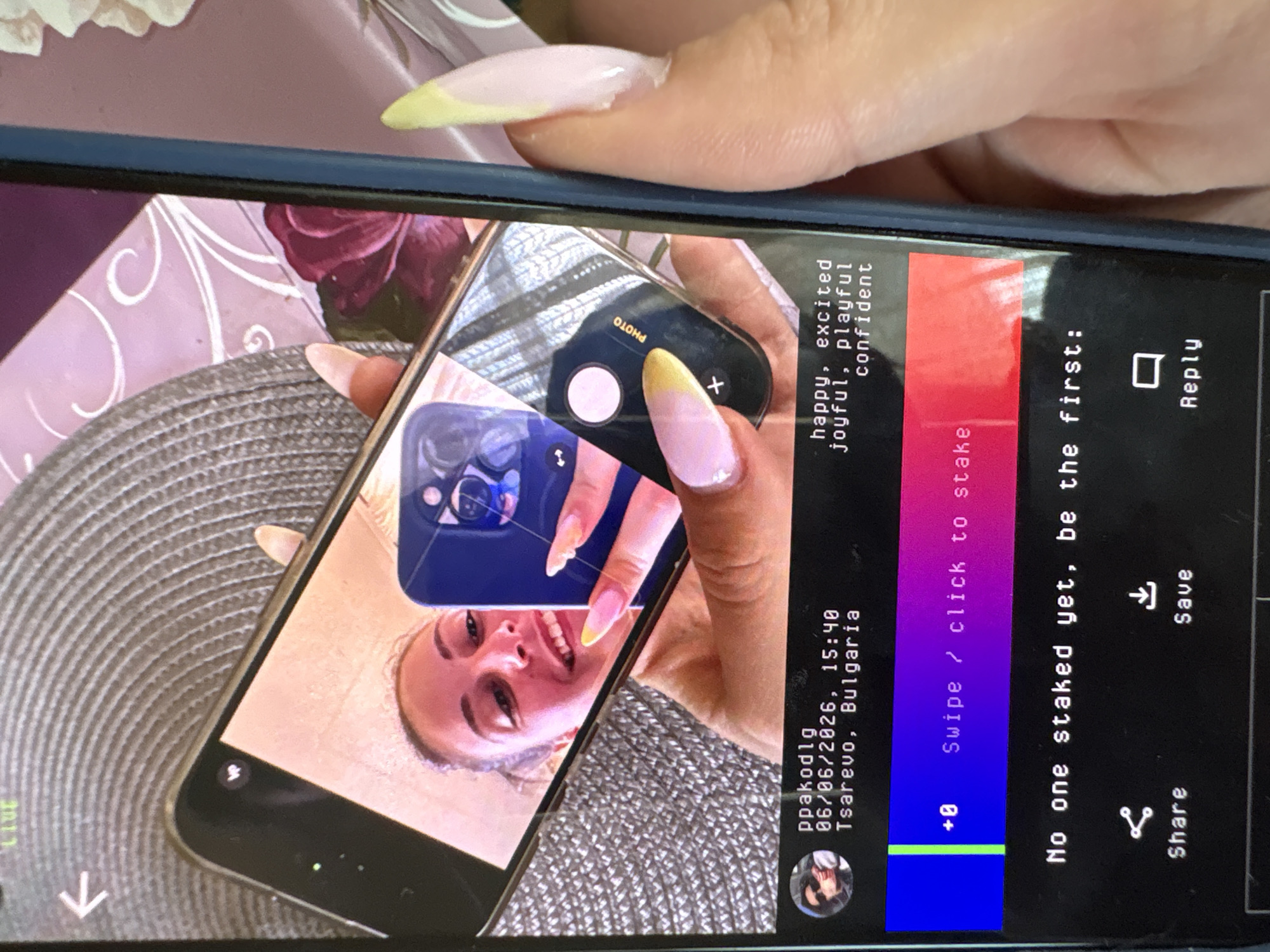 The image displays a photograph of a smartphone screen, which itself shows a digital post. A hand with long, light pink nails tipped with yellow is holding the smartphone on the right side of the frame.
The smartphone screen prominently features a selfie taken by a smiling woman. She is holding a dark blue smartphone, clearly visible in the selfie, with its camera interface displaying a white circular shutter button, "PHOTO" text, and a '+' icon. The woman's hand holding this inner smartphone also exhibits long, yellow-tipped manicured nails. The background within the selfie appears to be an interior wall of a light, warm hue.
Around the selfie on the displayed screen, a dark grey, textured, possibly woven circular placemat is visible on the left, and a pink surface adorned with white swirling patterns and a dark red rose is on the right.
Below the main image on the screen, a user interface is present:
* A circular profile picture shows a person wearing sunglasses and a hoodie.
* Next to it, the username "ppakodlg" is displayed.
* Below the username, the date and time "06/06/2026, 15:40" are specified, along with the precise location "Tsarevo, Bulgaria".
* A list of keywords reads "happy, excited joyful, playful confident".
* A horizontal bar with a gradient from blue to red, marked with a green vertical line and "+0", prompts "Swipe / click to stake".
* Further down, the text "No one staked yet, be the first:" encourages interaction.
* At the bottom, three icons with corresponding labels "Share", "Save", and "Reply" are visible.
The top left of the screen shows a white left-arrow icon and partially visible green text "club".