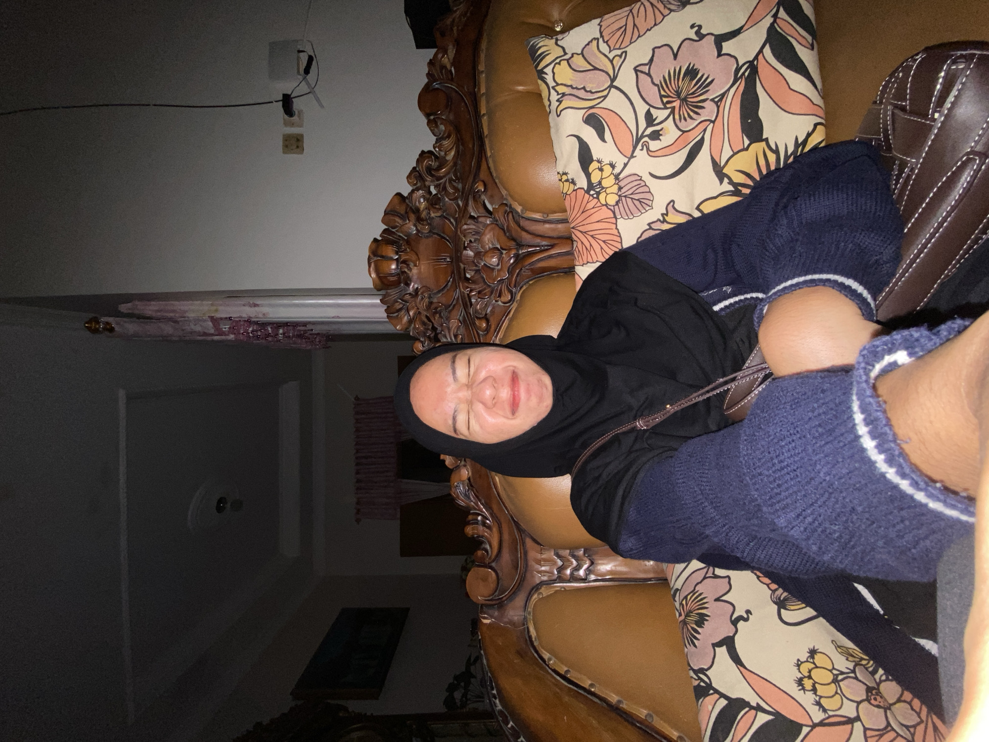 A young woman wearing a black hijab and a dark blue sweater sits on a brown leather sofa. She is facing the camera with her eyes closed and her face scrunched up, as if laughing or making a funny face. The sofa has ornate carved wood detailing on the back and is decorated with pillows featuring a floral pattern in shades of pink, yellow, and orange. The background is dimly lit, showing a ceiling with a light fixture and a portion of a wall with electrical outlets and a curtain. The scene appears to be indoors, possibly in a living room in Margorejo, Indonesia, though this location is not explicitly visible in the image.
