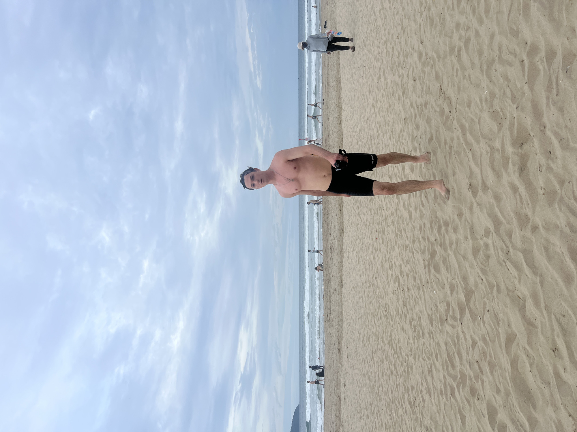 A man, shirtless and wearing black swim trunks, walks on a sandy beach with the ocean and waves in the background. The sky is overcast. Other people are visible in the distance, some wading in the water and others on the sand. The scene is likely My Khe Beach in Da Nang, Vietnam, a popular tourist destination known for its long stretches of sand and calm waters.
