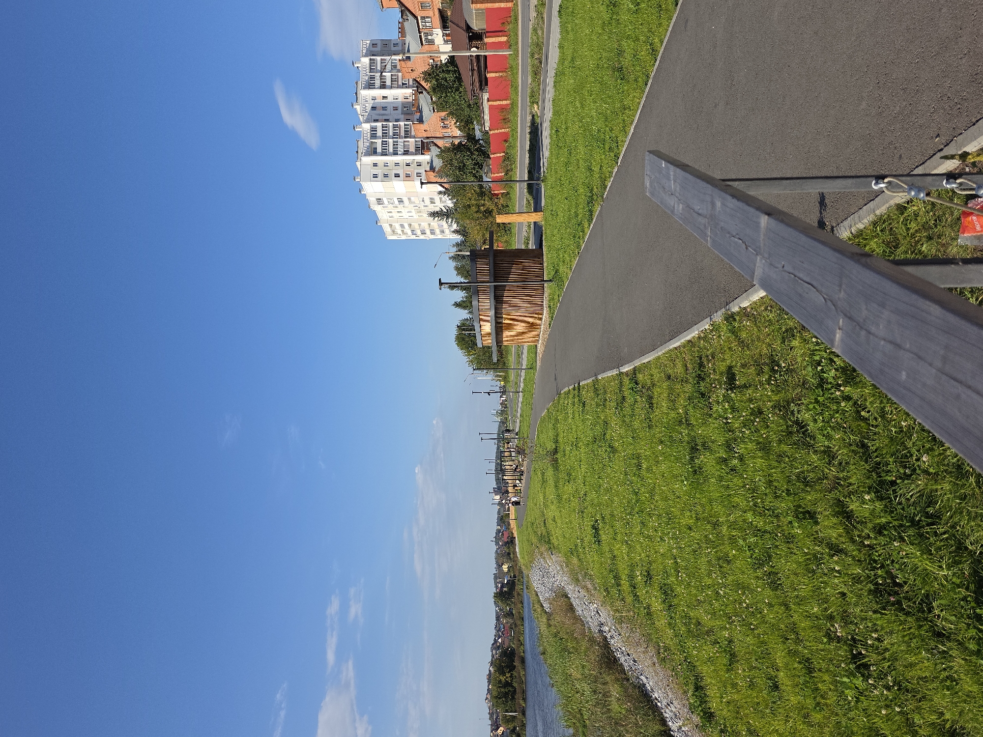 The image depicts a paved walkway with a grassy area on the left bordering a body of water, likely a river or lake. In the distance, a town or city skyline with residential buildings and trees is visible. The walkway features streetlights and a small wooden pavilion-like structure. A low wooden railing is in the foreground. The sky is clear blue with a few scattered clouds. The location is identified as the Embankment of the River Miass in Miass, Russia.