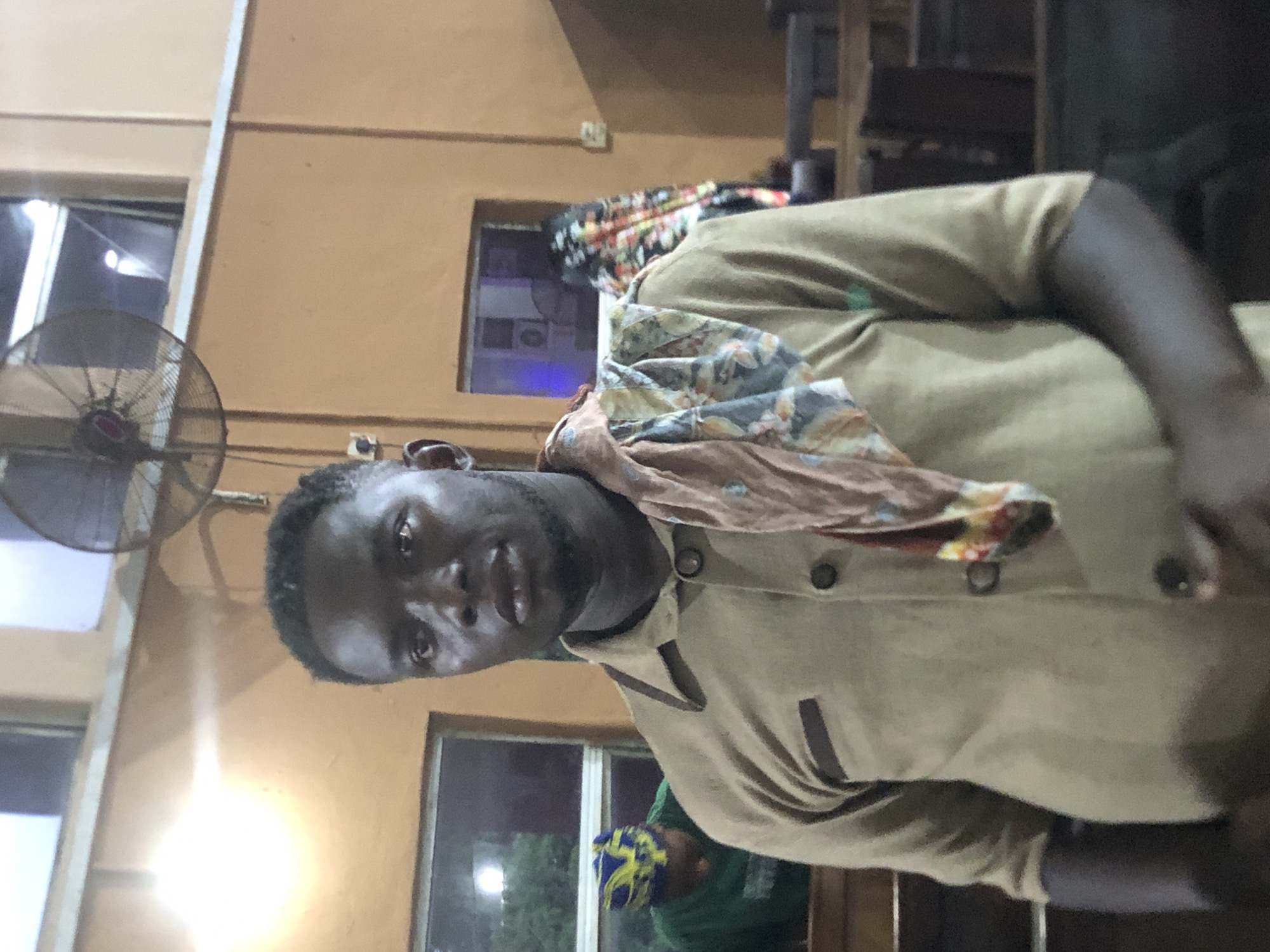 A man stands indoors in Nsugbe, Nigeria. He has a scarf draped over his left shoulder. He wears a collared shirt with buttons and dark trim. In the background is an electric fan mounted to the wall and window views of outdoors at night.