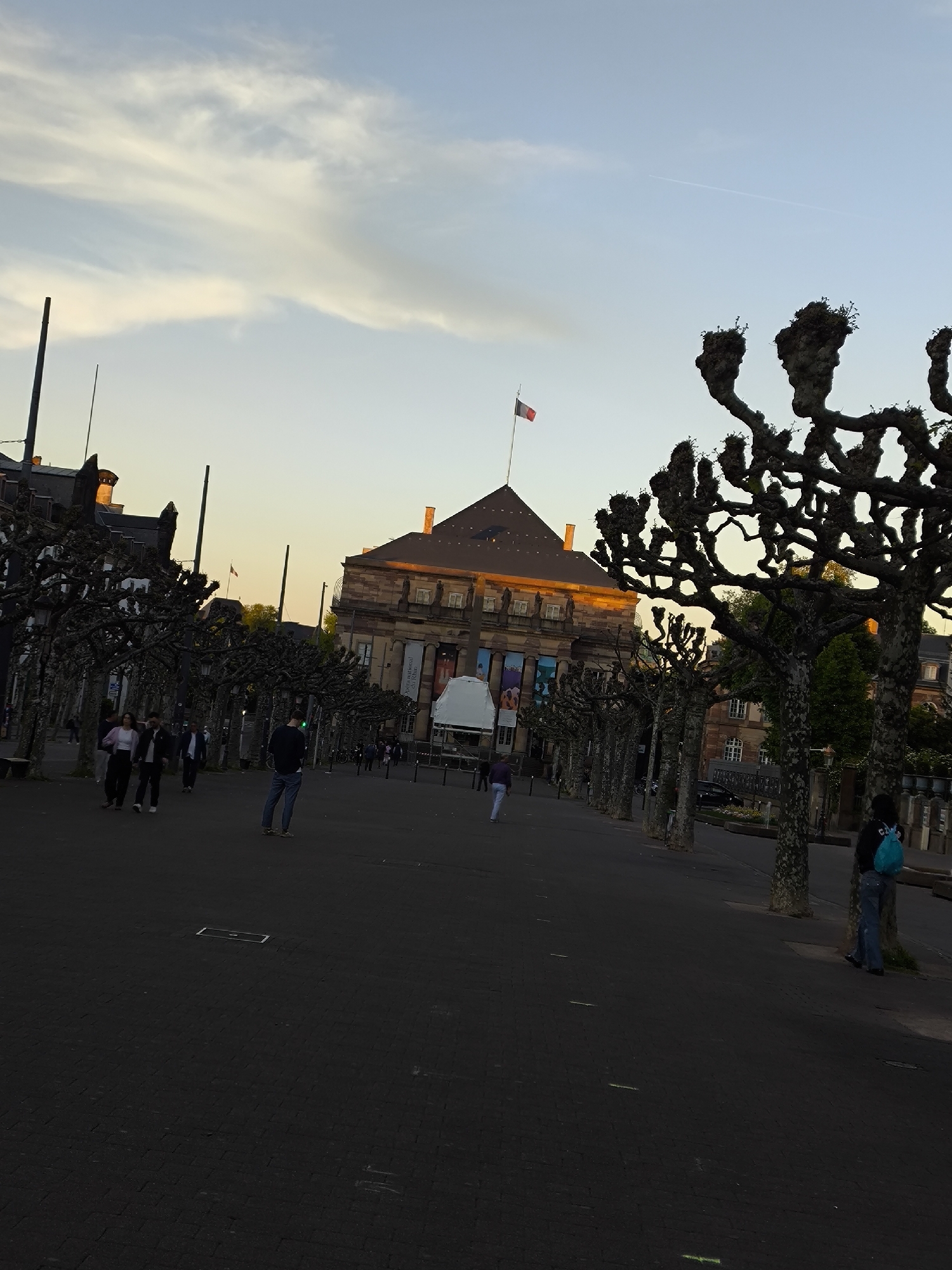 The image depicts a public square in Strasbourg, France, with the Palais Rohan visible in the background. The building is a large, ornate structure with a red-and-white French flag atop its roof. In front of the Palais Rohan, a white tent-like structure is set up. The square is paved with dark-colored bricks and lined with rows of bare trees with distinctive, knobby branches. Several people are present in the square: some are walking along the main path, while a person wearing a blue backpack is standing near a tree on the right side of the frame. The sky above is light blue with streaks of white clouds, suggesting either early morning or late afternoon light. The overall impression is one of a public space with a mix of historical architecture and contemporary activity.