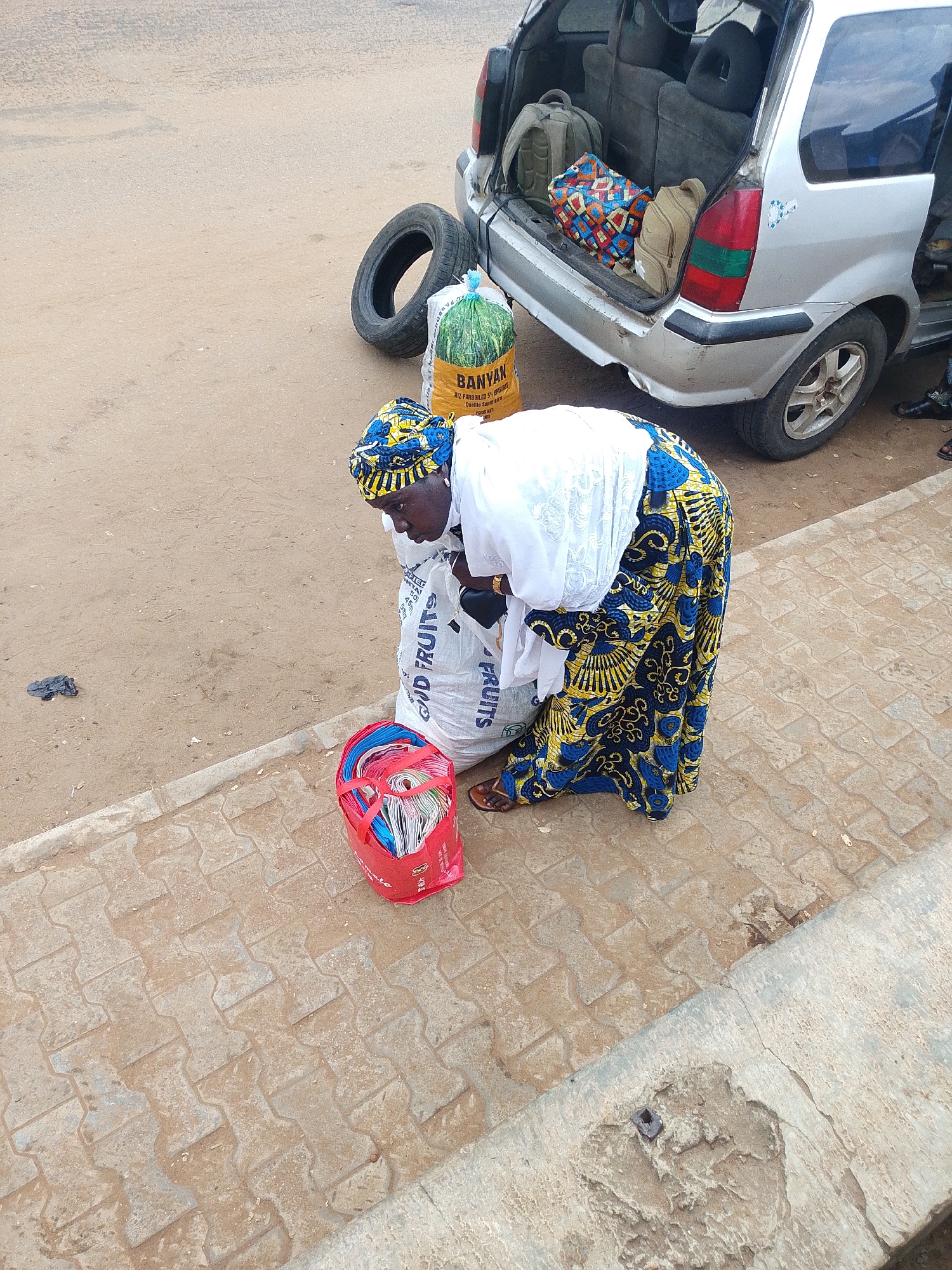 A woman, dressed in a white top and a vibrantly patterned blue and yellow skirt and headscarf, is engaged in an activity near the open back of a silver van. She is bent over, partially obscured by a large white sack. Beside her, also on the ground, is a red plastic bag filled with what appear to be folded items, possibly clothes or fabric. Another large sack, green and yellow, with the word "BANYAN" prominently displayed on it, lies on the ground next to a spare tire. The van's interior is visible, containing several bags and backpacks. The scene takes place on a paved walkway next to a dusty road. The lighting suggests it is daytime. The overall impression is of a commercial transaction or loading/unloading of goods.