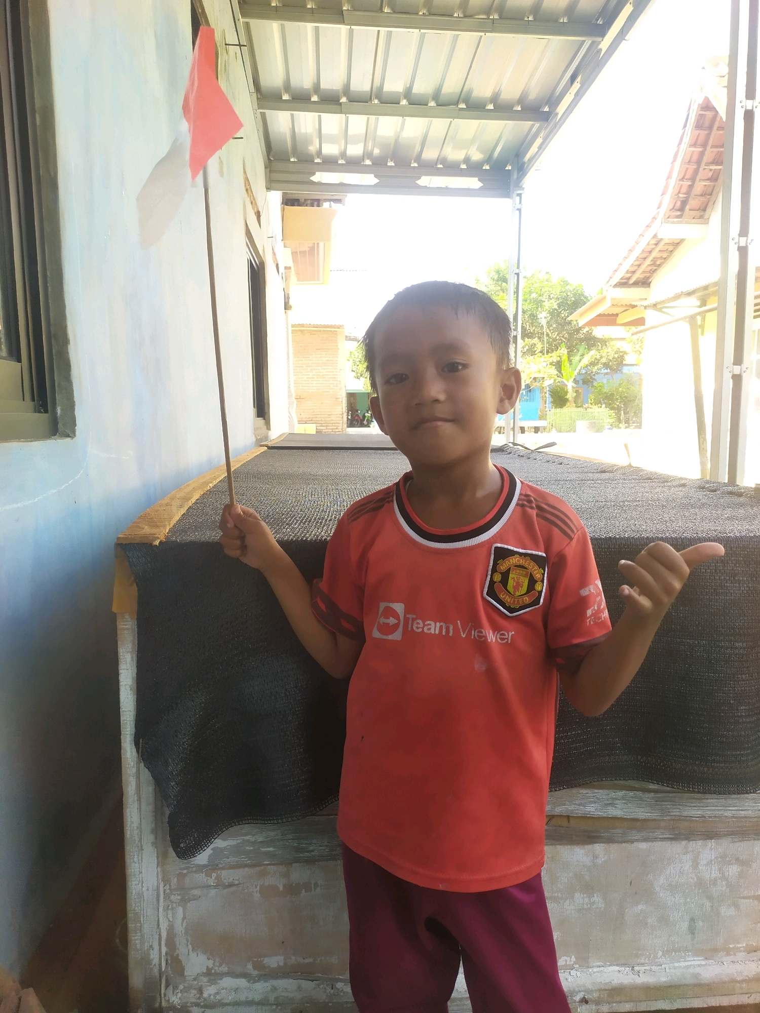 A young boy stands outdoors, holding a small red flag on a stick. He is wearing a red t-shirt with the Manchester United logo and the words "Team Viewer" printed on it, and dark red pants. The boy is standing in front of a large, dark-colored, rectangular structure covered with a mesh material. Behind the structure, a building with a tiled roof is visible, along with trees and other vegetation. The scene appears to be in a semi-rural or village setting, possibly in Desa Penggalang, Kecamatan Adipala, Indonesia, given the architectural style and vegetation. The lighting suggests it is daytime, likely sunny. The boy is looking towards the camera.