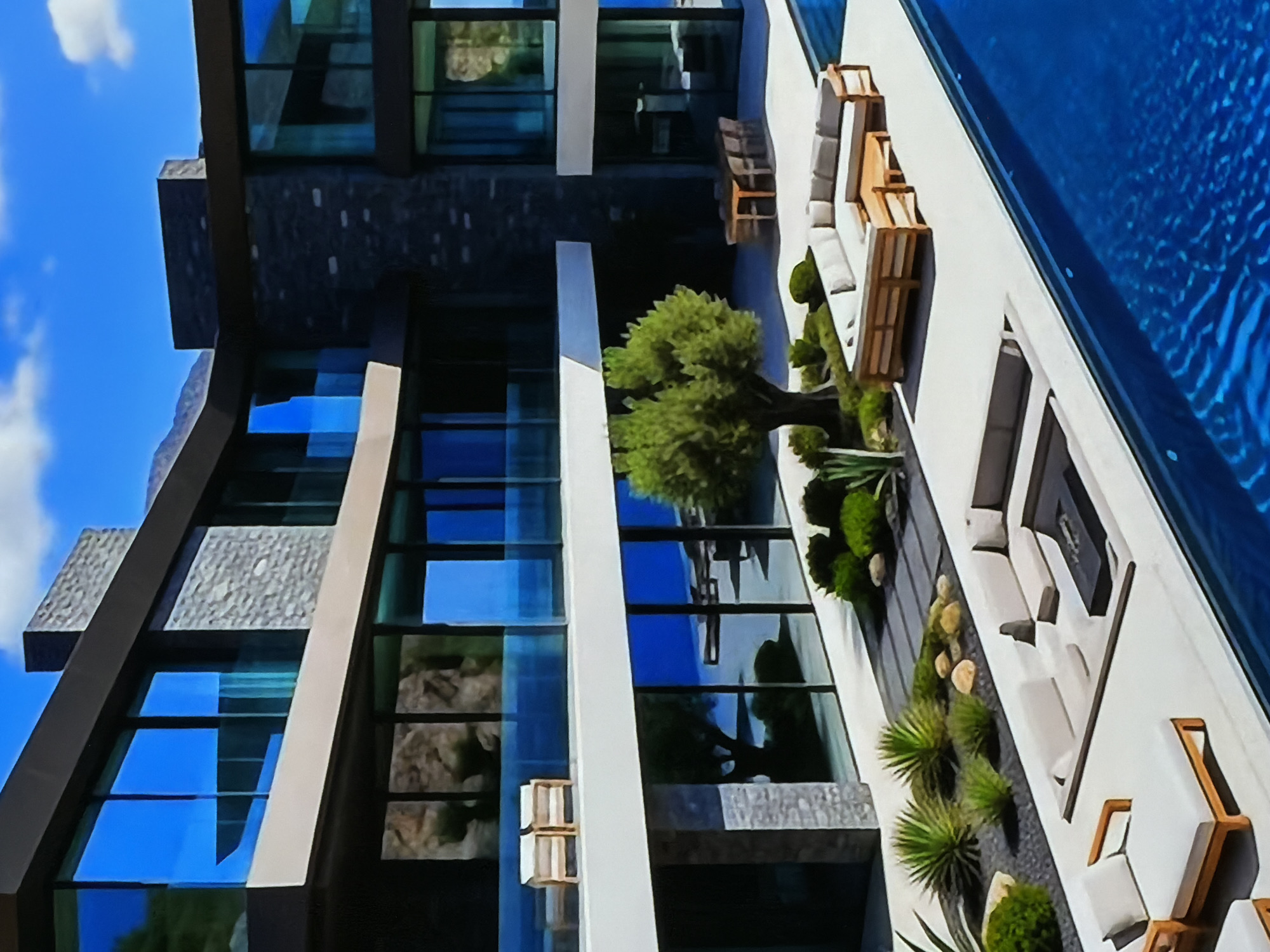 This image depicts a modern, luxurious residential property under a clear, bright blue sky with sparse clouds. The multi-story building showcases contemporary architecture, characterized by large, reflective glass windows framed in dark materials and interspersed with gray stone accents. The windows extensively reflect the sky, clouds, and nearby foliage, blending the structure with its environment.
An expansive outdoor living area adjoins the building. A pristine, rectangular swimming pool with deep blue, rippled water stretches parallel to the residence. The patio is meticulously landscaped, featuring a variety of green plants, including a central deciduous tree, manicured shrubs, and spiky succulents, arranged among decorative light-colored rocks. Modern outdoor furniture is strategically placed, including sofas, chairs with light cushions, and wooden loungers, suggesting multiple zones for relaxation.
The scene is tranquil and devoid of people, implying an unoccupied moment. The bright lighting indicates a sunny daytime setting. The overall impression is one of sophisticated design, comfort, and serenity within a high-end property. The provided location context is Sofiivska Borshchahivka, Ukraine. There is no visible text in the image.